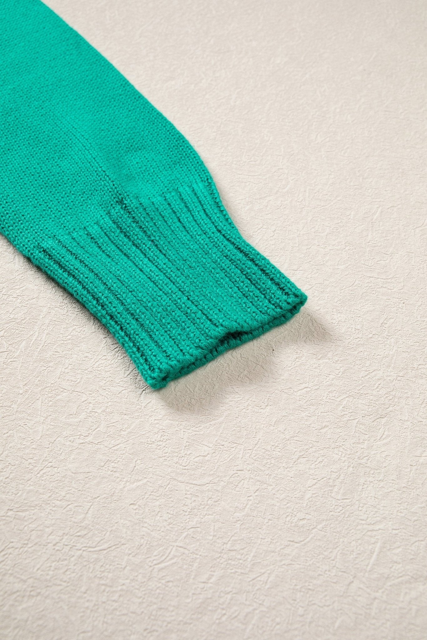 A close-up of a bright teal green ribbed knit sleeve cuff on a textured off-white background.