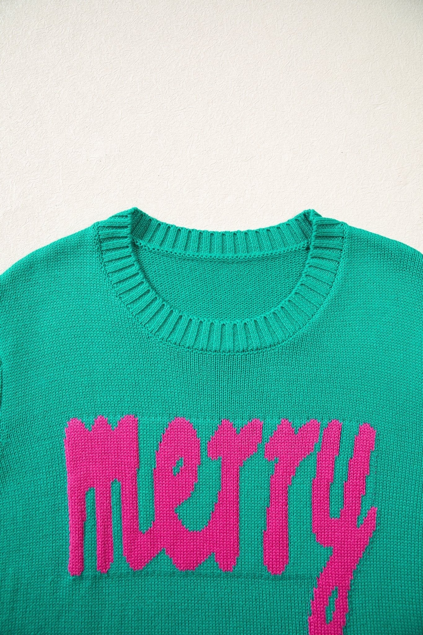 Close-up of bright green knit sweater with “merry” in bright pink across the chest on an off-white background.