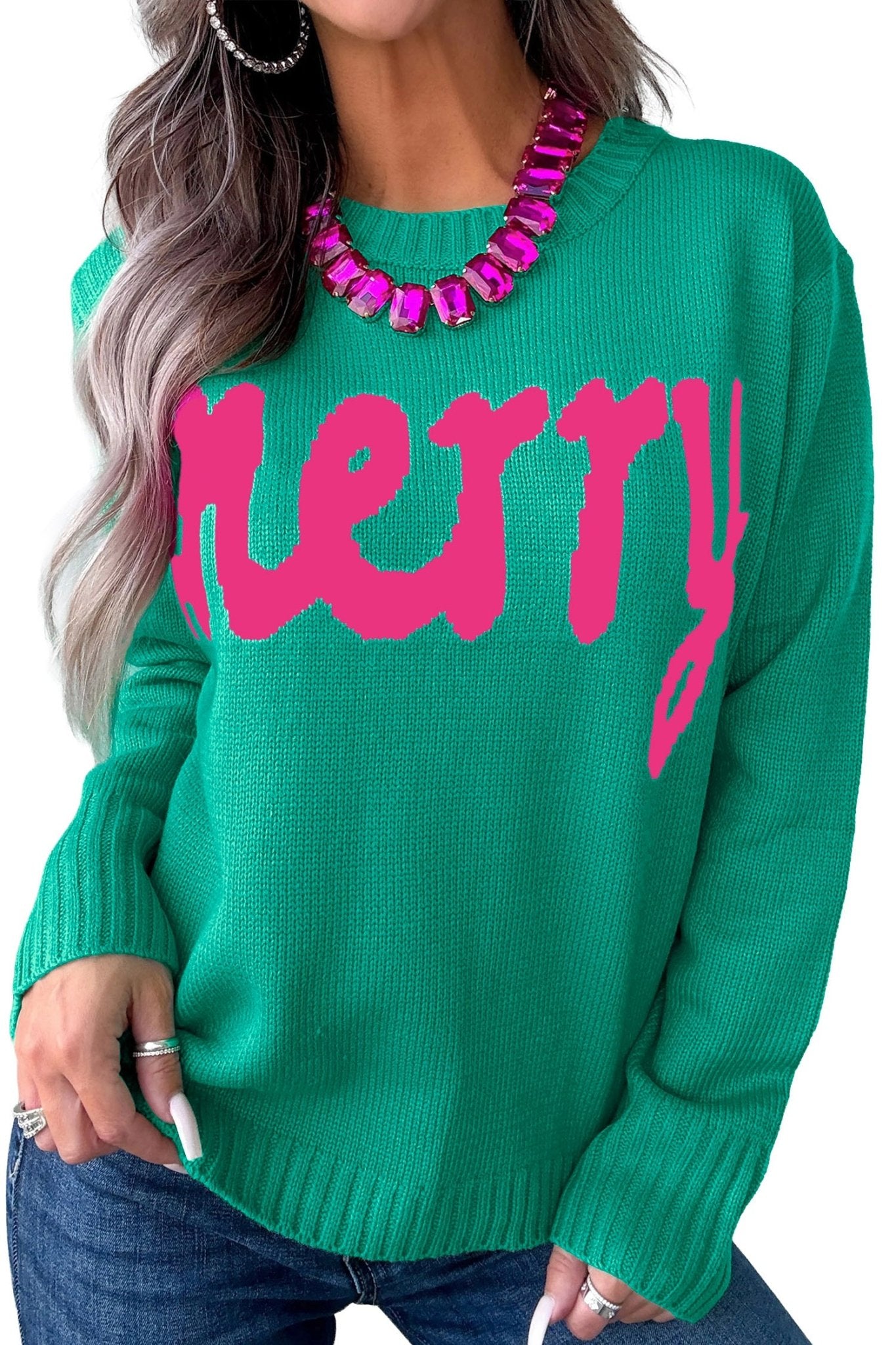Woman wearing bright green “Merry” knit sweater with blue jeans & chunky pink statement necklace.