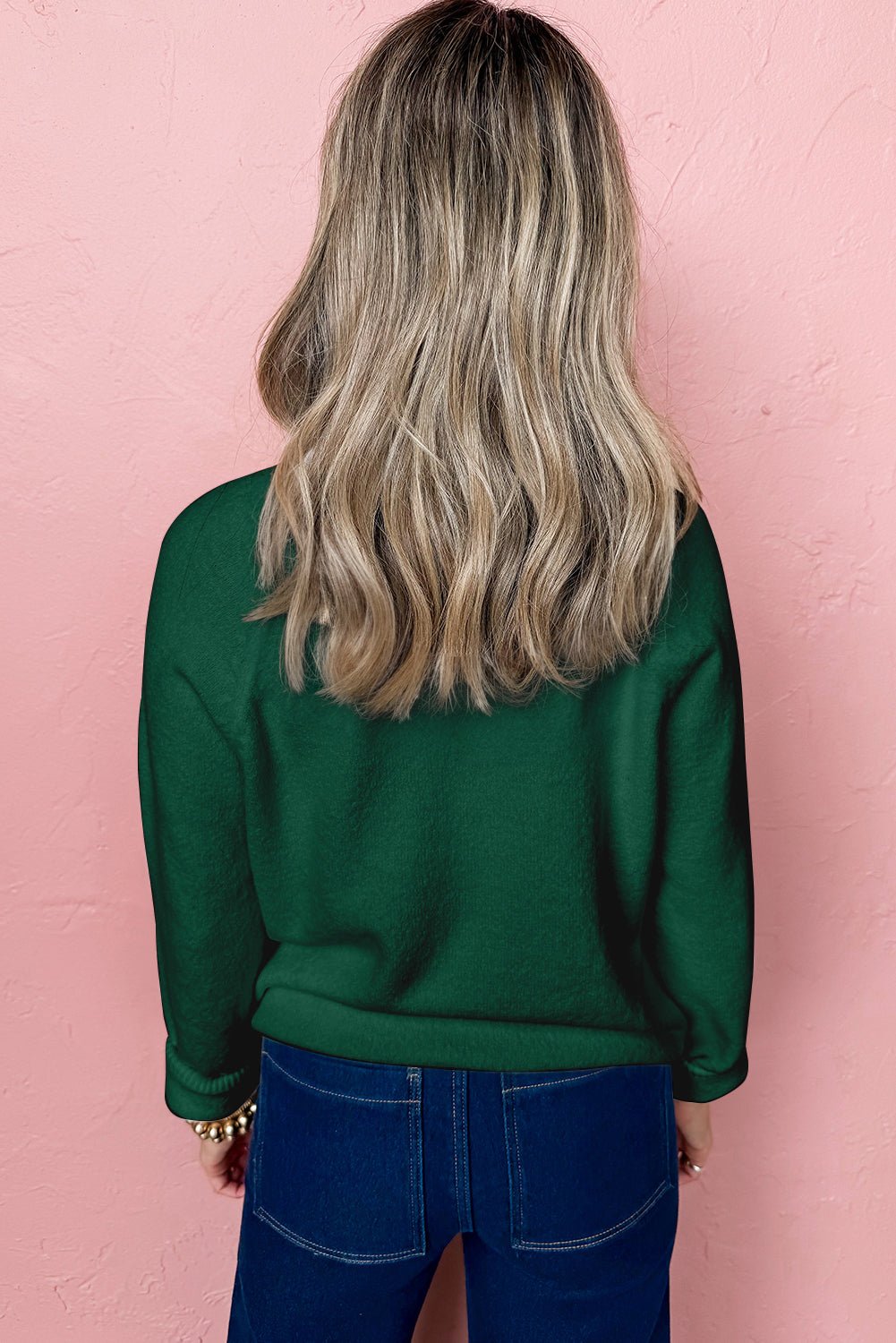 Person wearing a green sweater and blue jeans against a pink wall