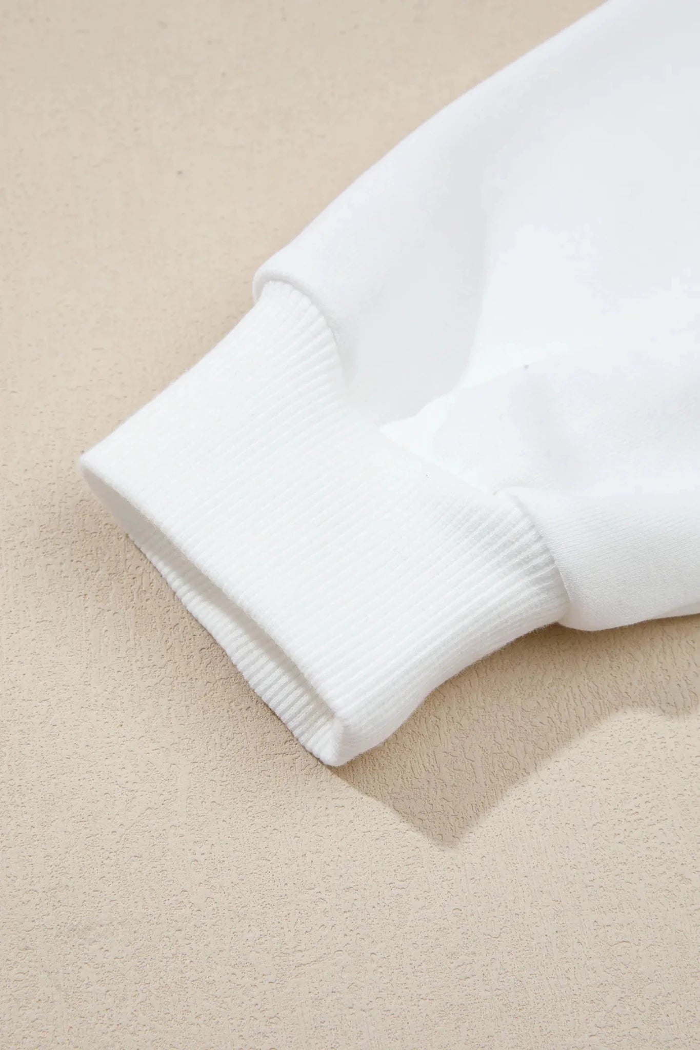 Close-up of a white ribbed knit cuff on a sweatshirt or sweater, laid on a textured beige surface.