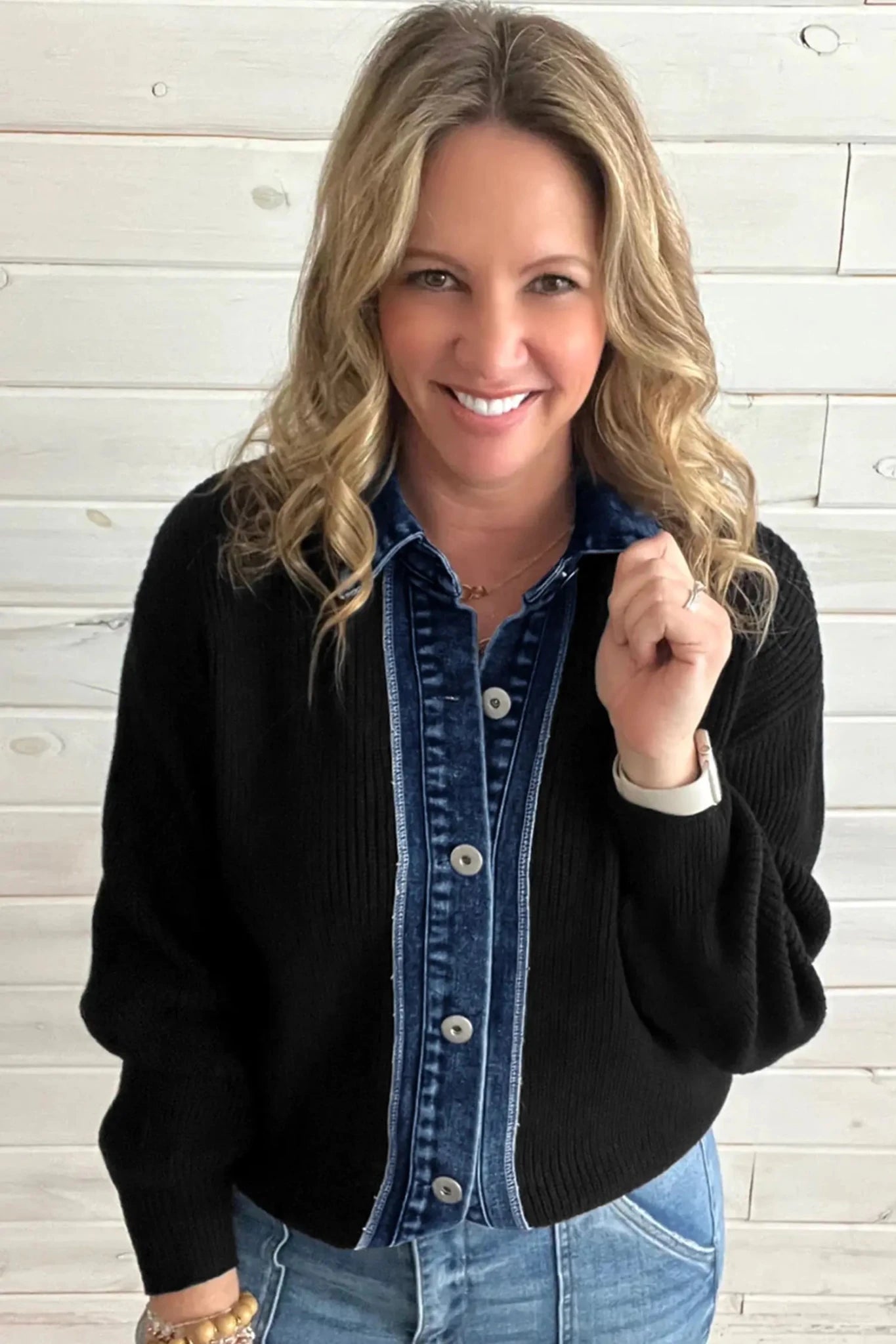 Smiling blonde woman in black ribbed cardigan over a blue denim shirt with light blue jeans against a white wood plank wall.