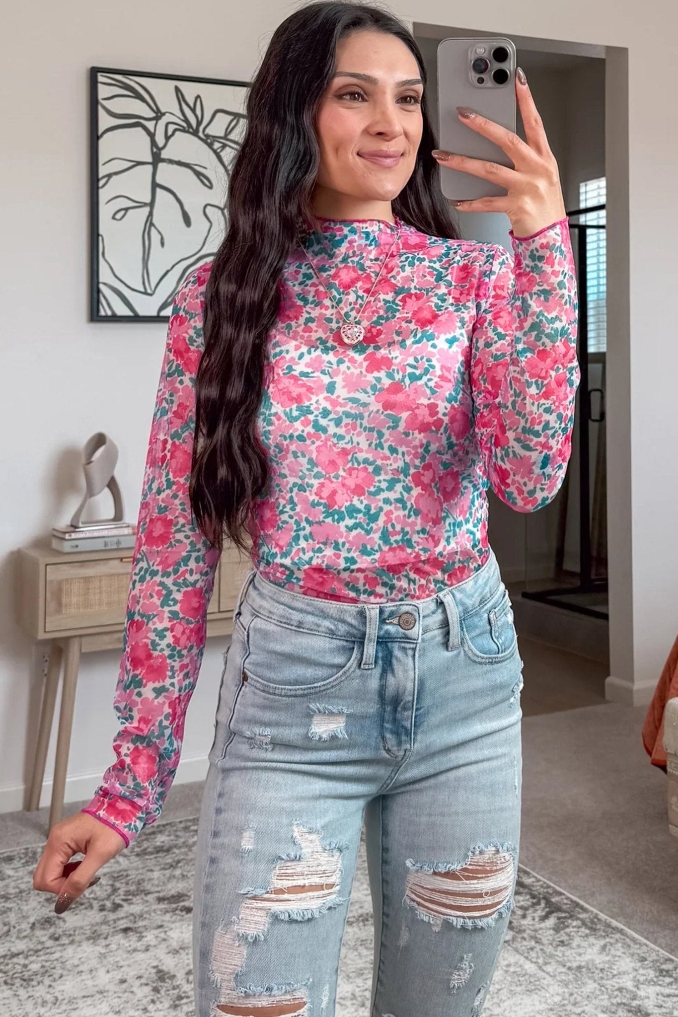 Woman taking a mirror selfie wearing a floral top and ripped jeans in a home setting.