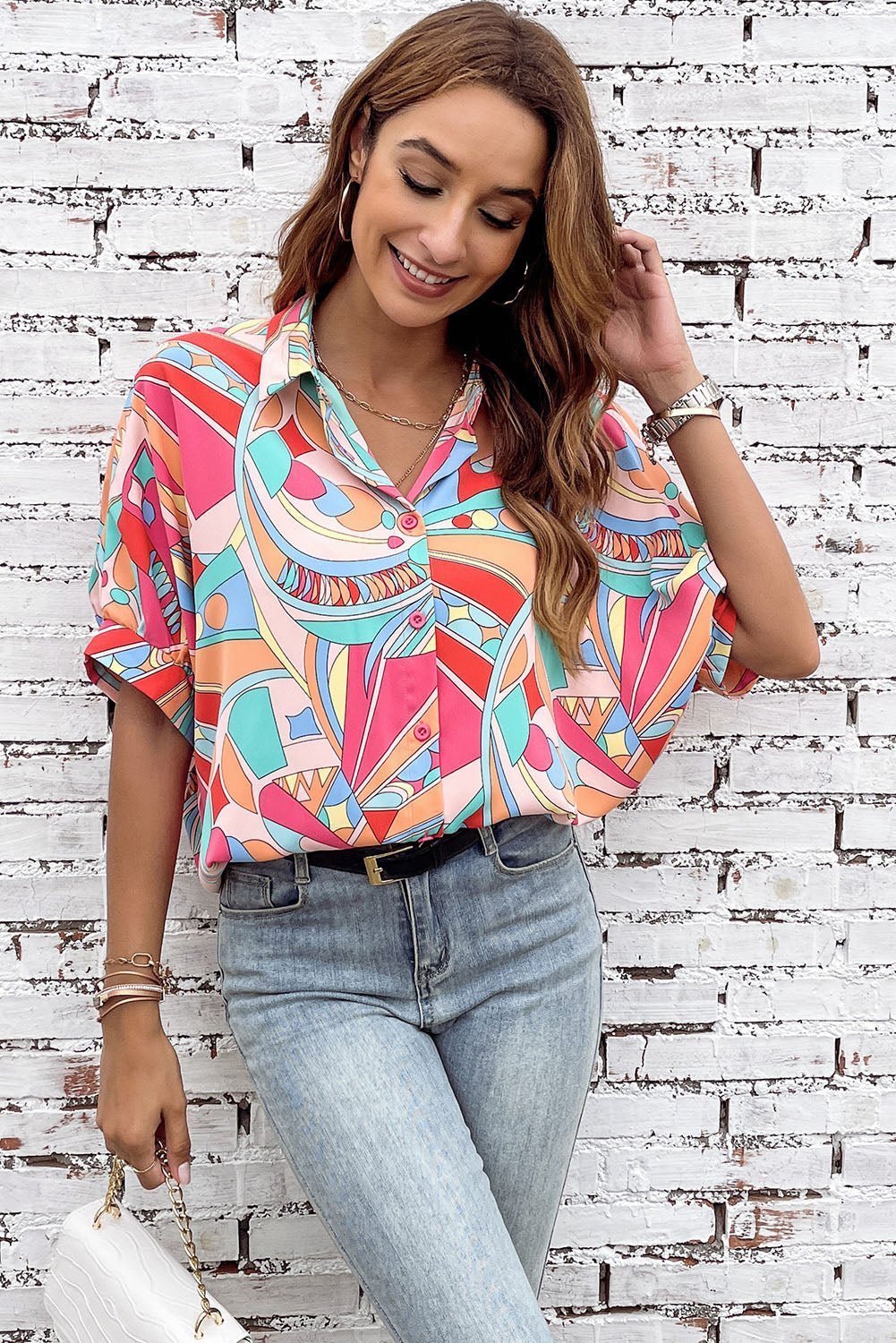 Woman wearing a colorful patterned blouse and light blue jeans against a white brick wall.
