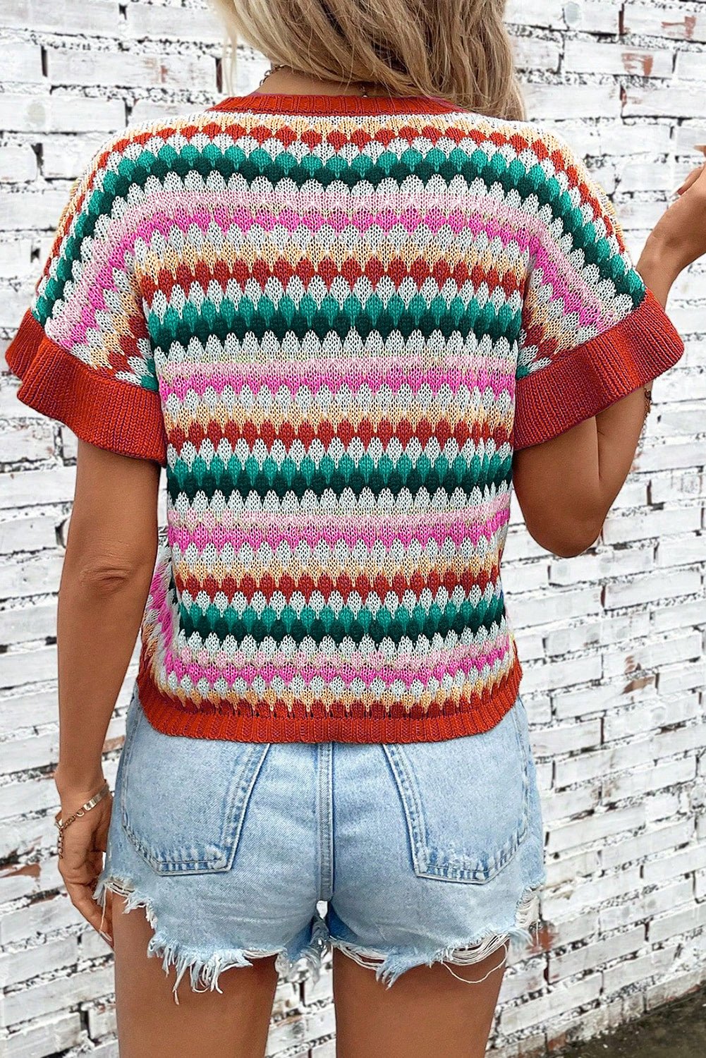 Person wearing a colorful striped sweater and denim shorts against a white brick wall.