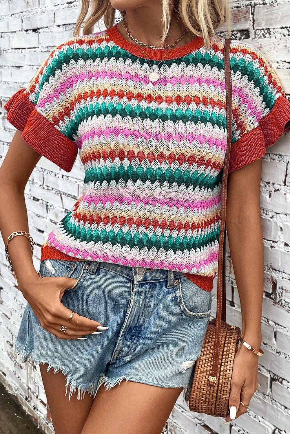 Person wearing a colorful striped sweater and denim shorts against a white brick wall.