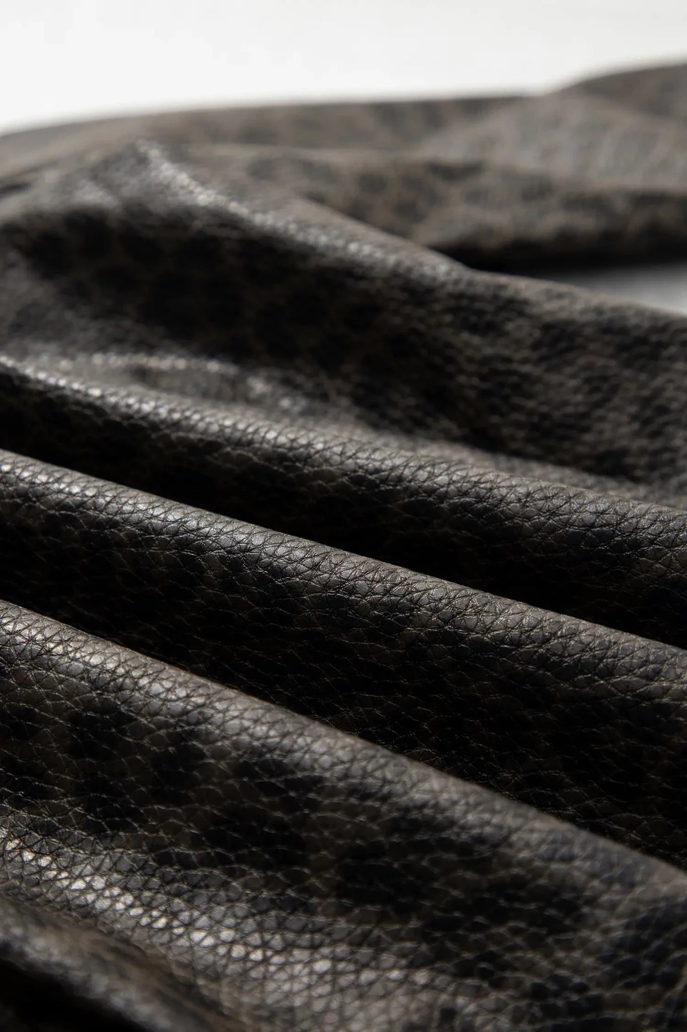 Close-up of dark textured leather with a pebbled grain and soft folds, photographed against a light neutral background.