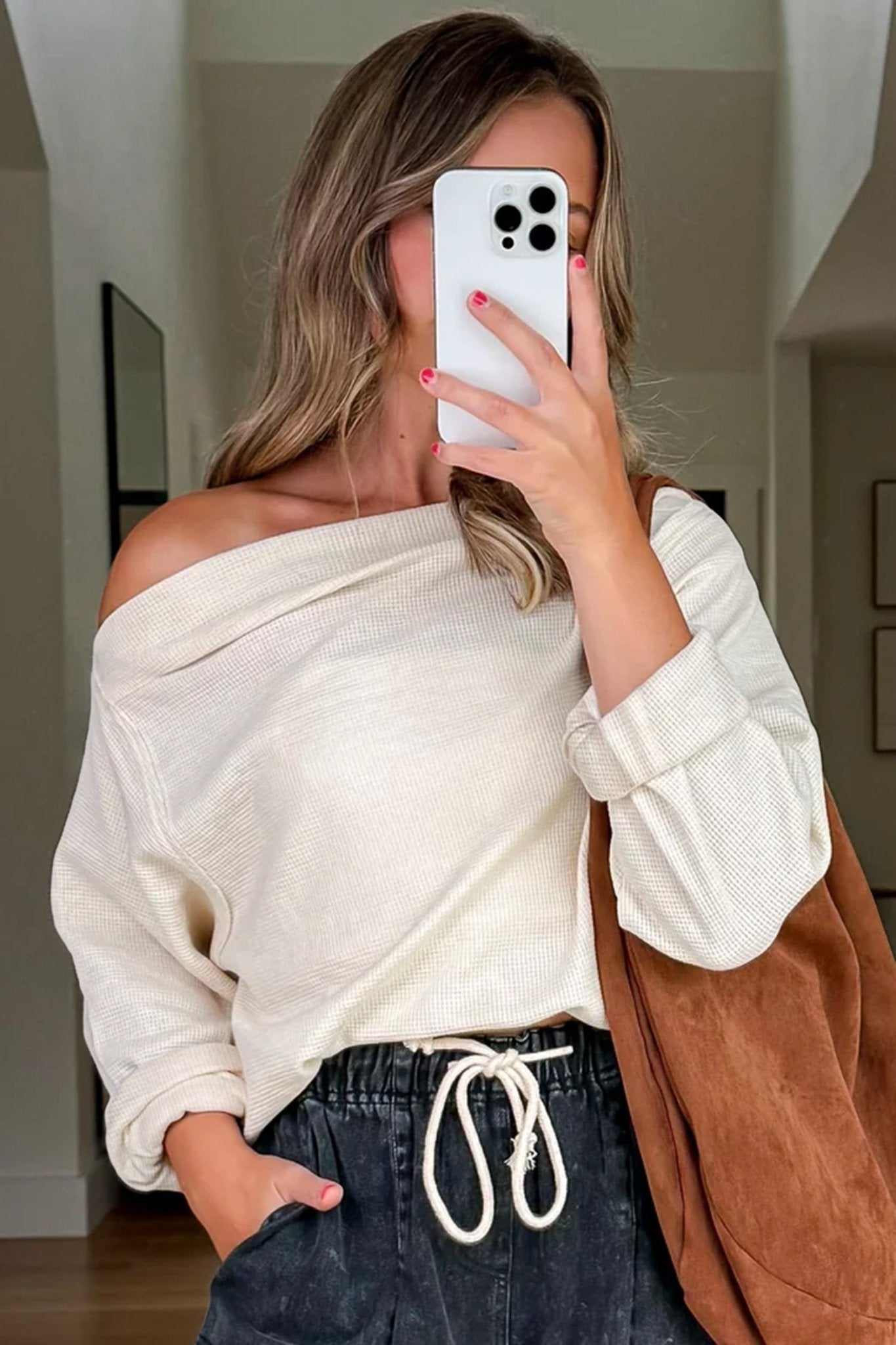 Woman taking a mirror selfie wearing an off-shoulder white sweater and dark pants.