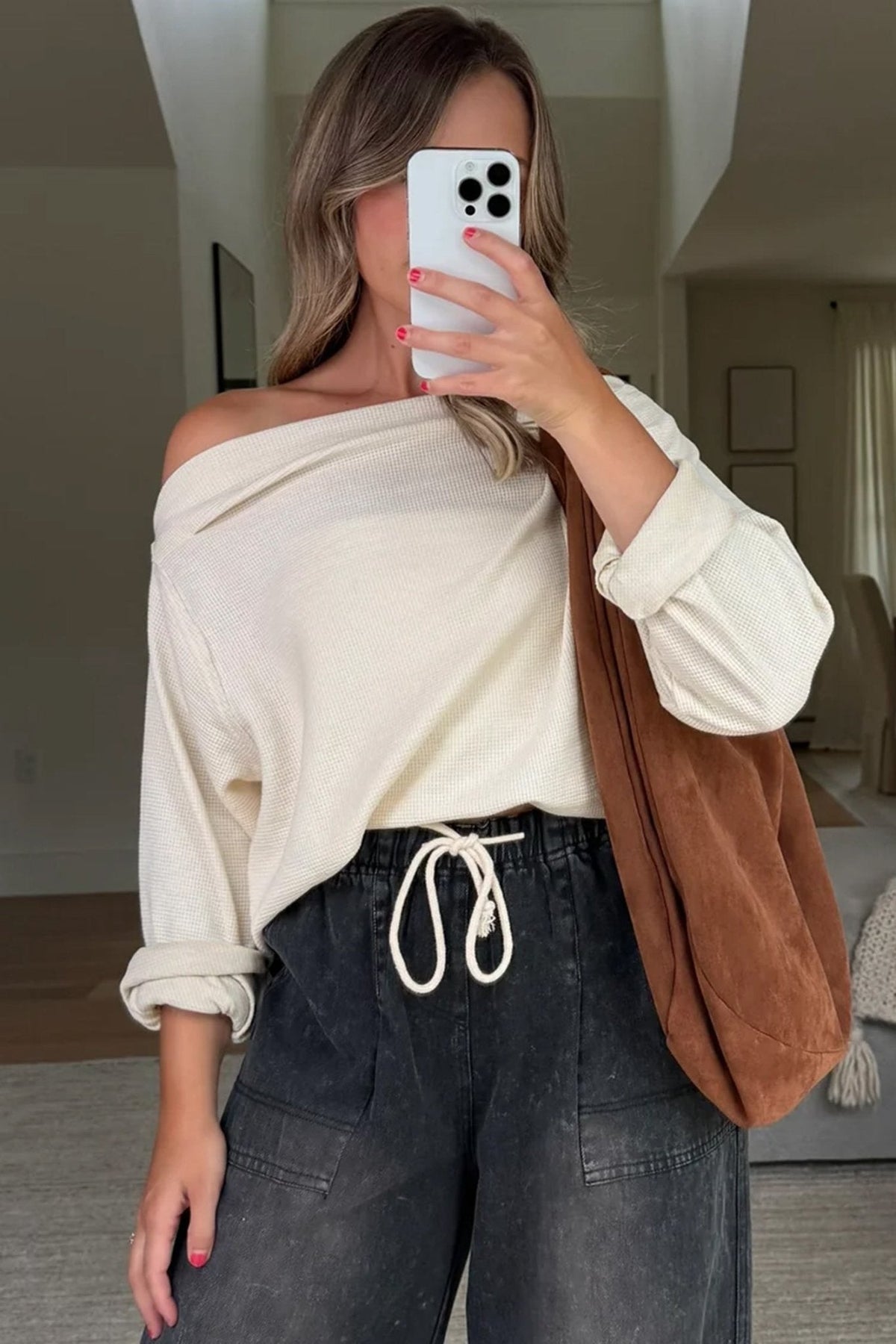 Woman taking a mirror selfie wearing a white off-shoulder top, dark jeans, and a brown bag.