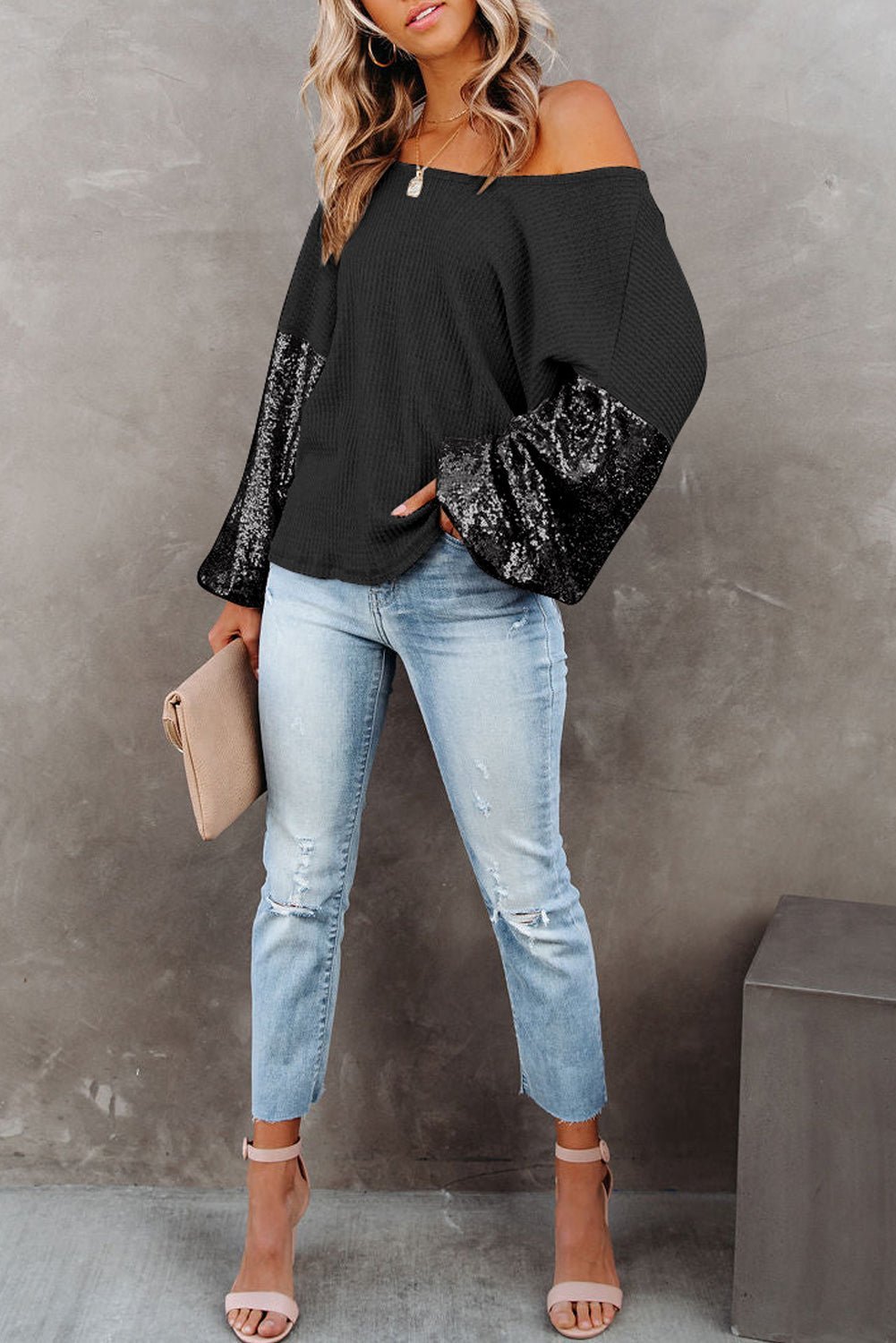 Woman wearing a black off-shoulder top with sequin sleeves and light blue jeans against a gray background.