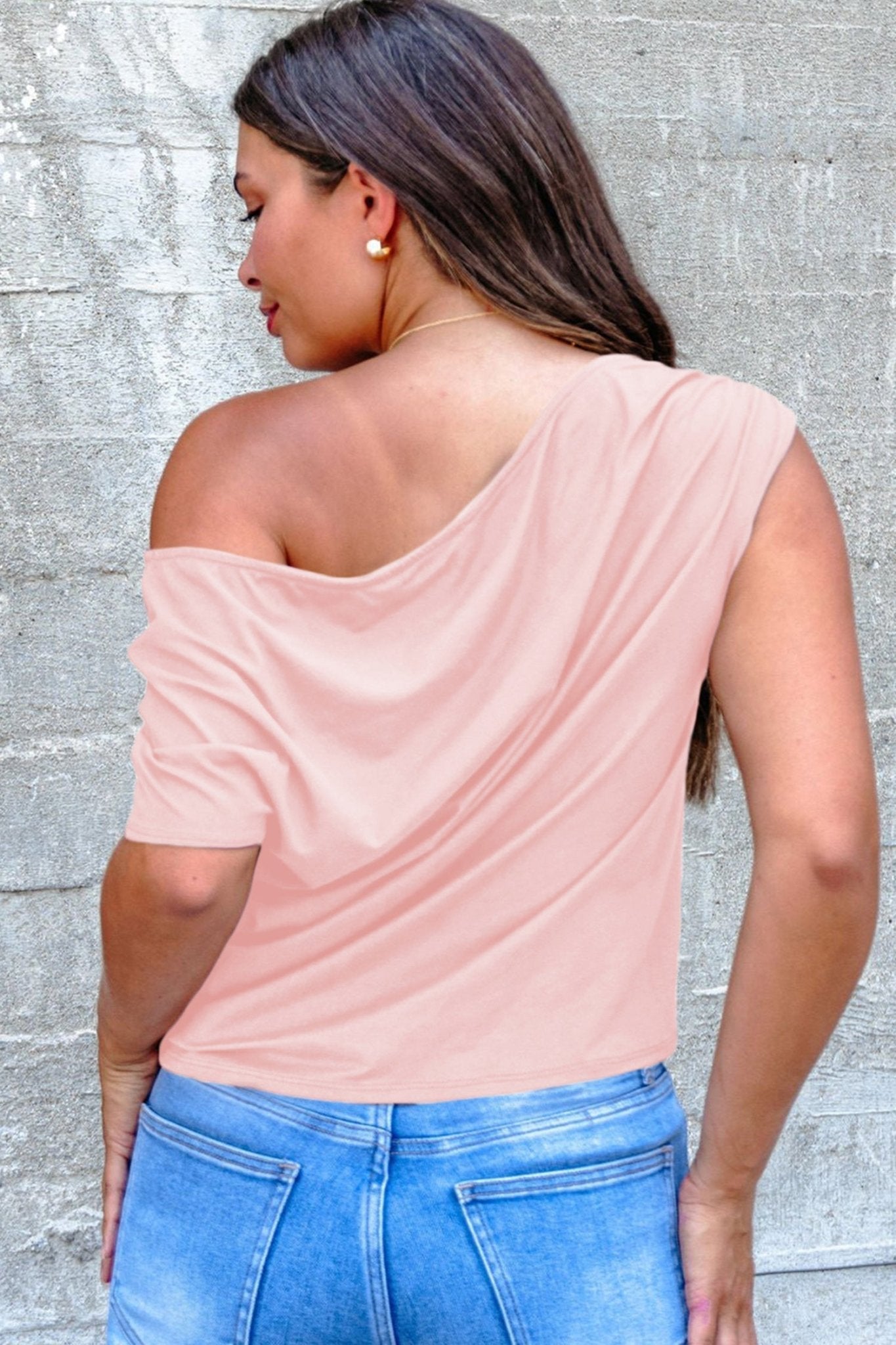 Woman wearing a pink off-shoulder top and blue jeans against a gray concrete wall.