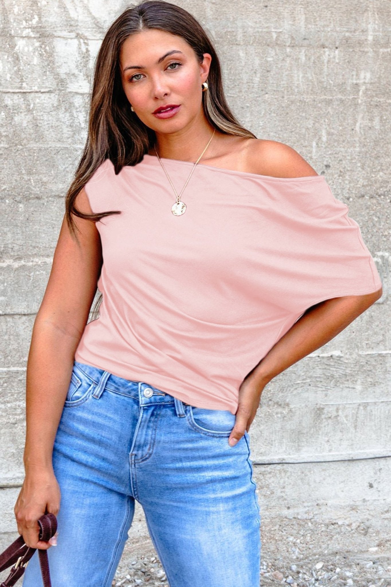 Woman wearing a pink off-shoulder top and blue jeans against a gray wall.