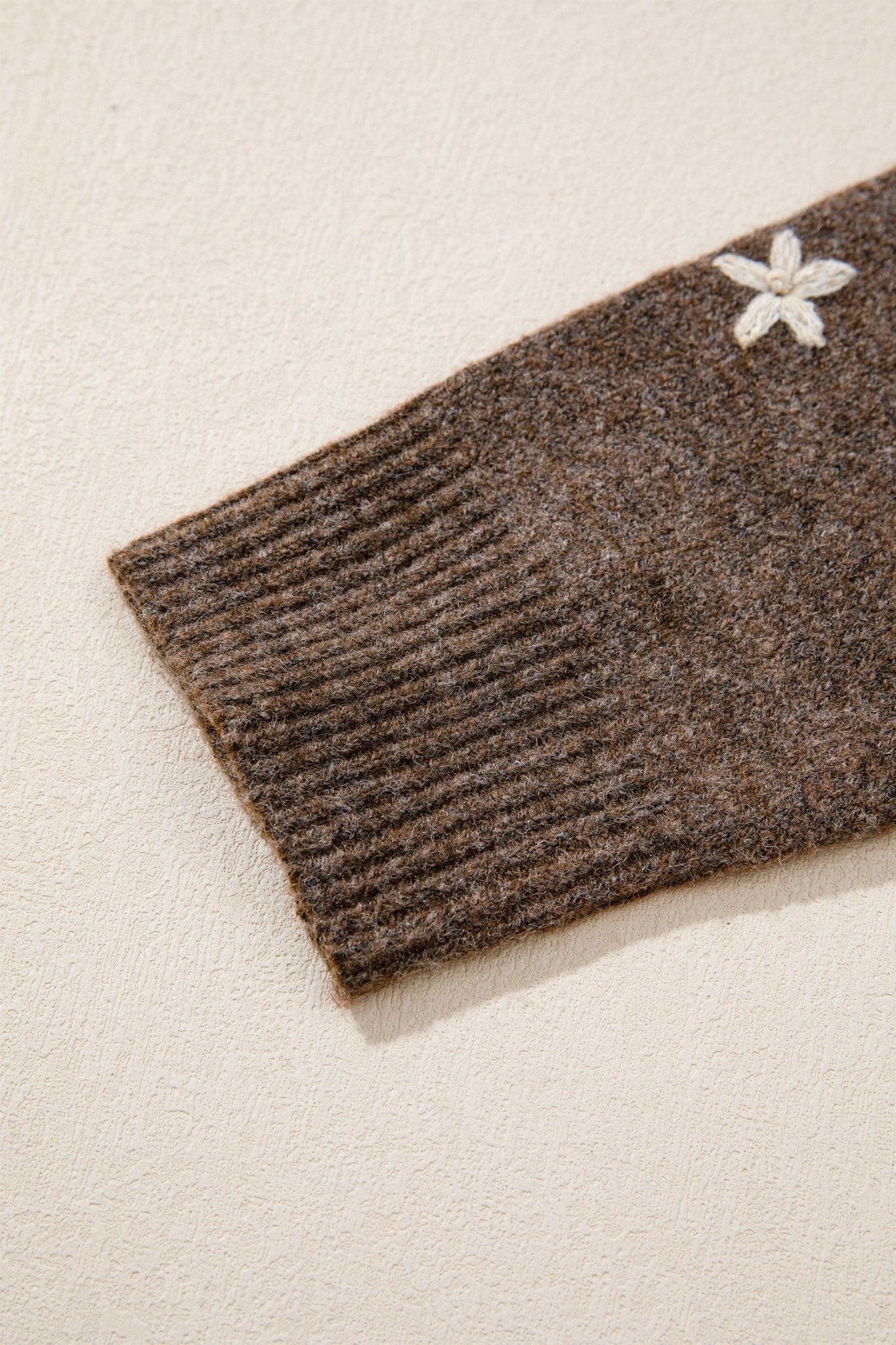 Close-up of a ribbed brown knit sleeve with a small white embroidered flower detail, set against a warm off-white textured background.