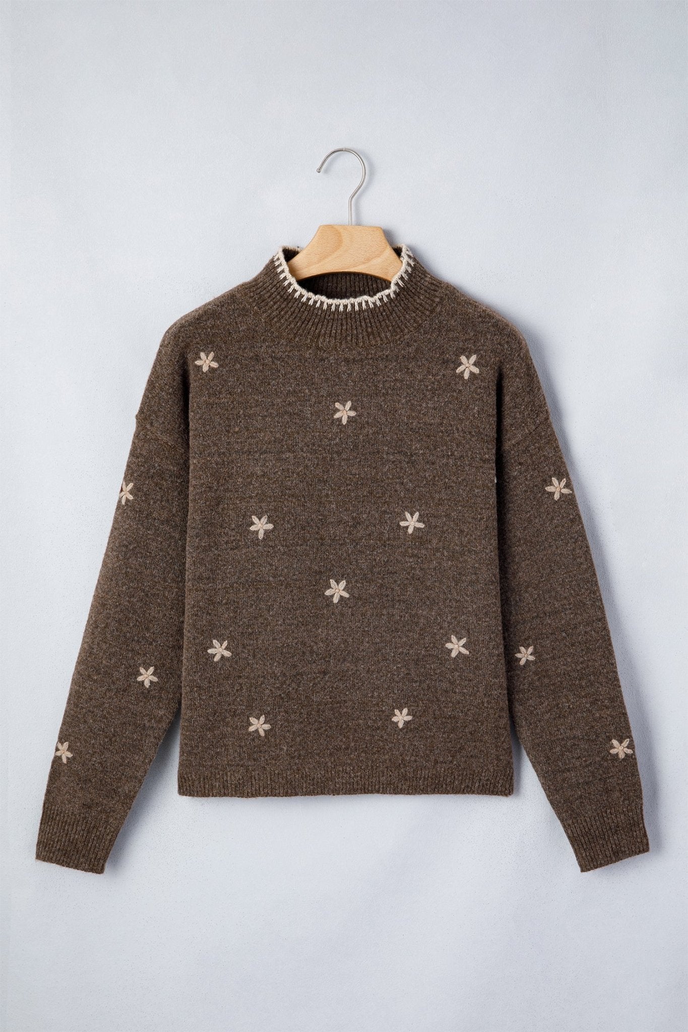 A brown knit sweater with small beige snowflake patterns, hanging on a wooden hanger against a light gray background.
