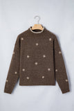 A brown knit sweater with small beige snowflake patterns, hanging on a wooden hanger against a light gray background.