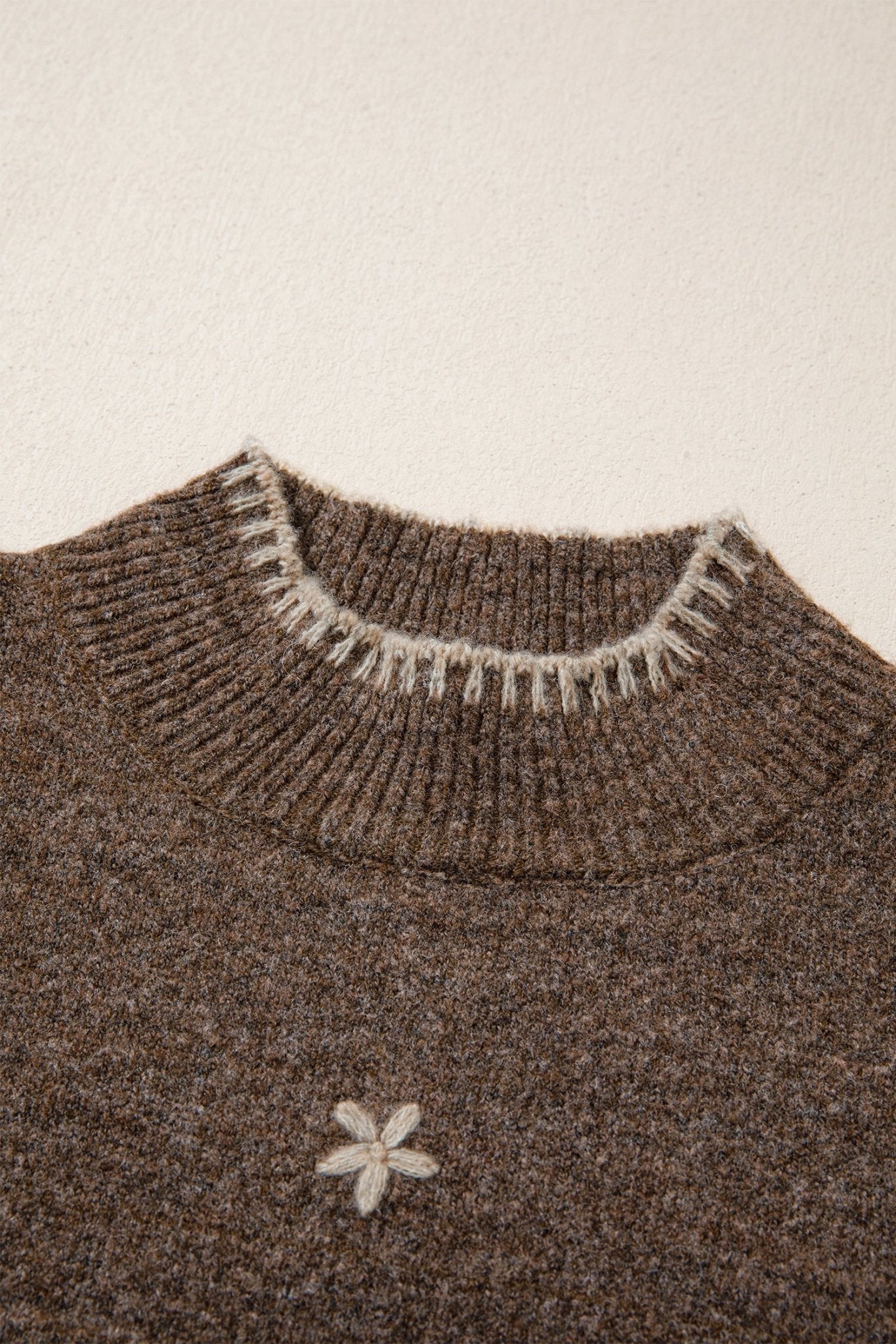 Close-up of a brown knit sweater with a ribbed crew neck featuring white fringe trim and a small white embroidered flower on the chest.