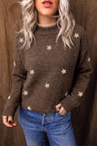 A woman wearing a brown knit sweater with small white floral motifs and a beaded mock neck, paired with blue jeans, standing against a brown textured backdrop.