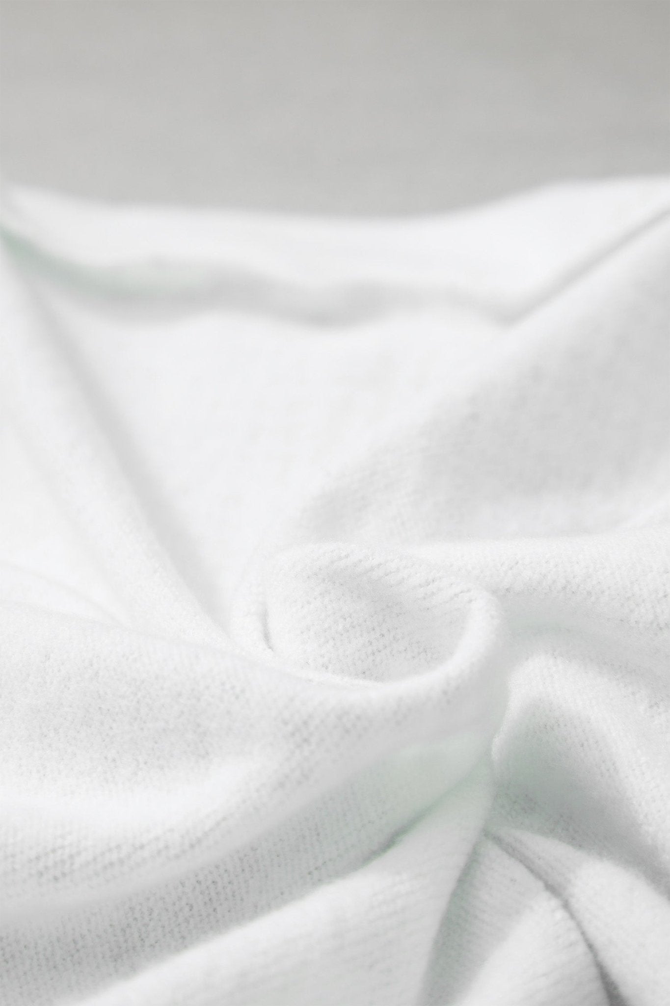 A close-up of soft white fabric with a fine knit texture, softly draped and folded against a light neutral background.