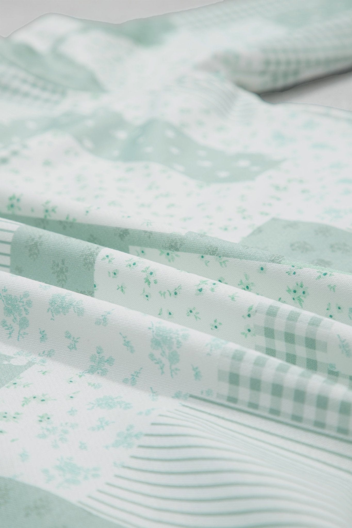 A close-up of folded fabric with a soft mint green and white floral print, gingham check, and striped pattern.