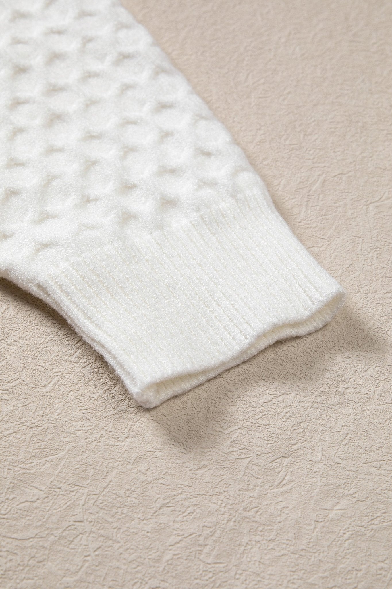 Close-up of a white ribbed knit sleeve or cuff with a textured waffle pattern, laid on a beige textured surface.