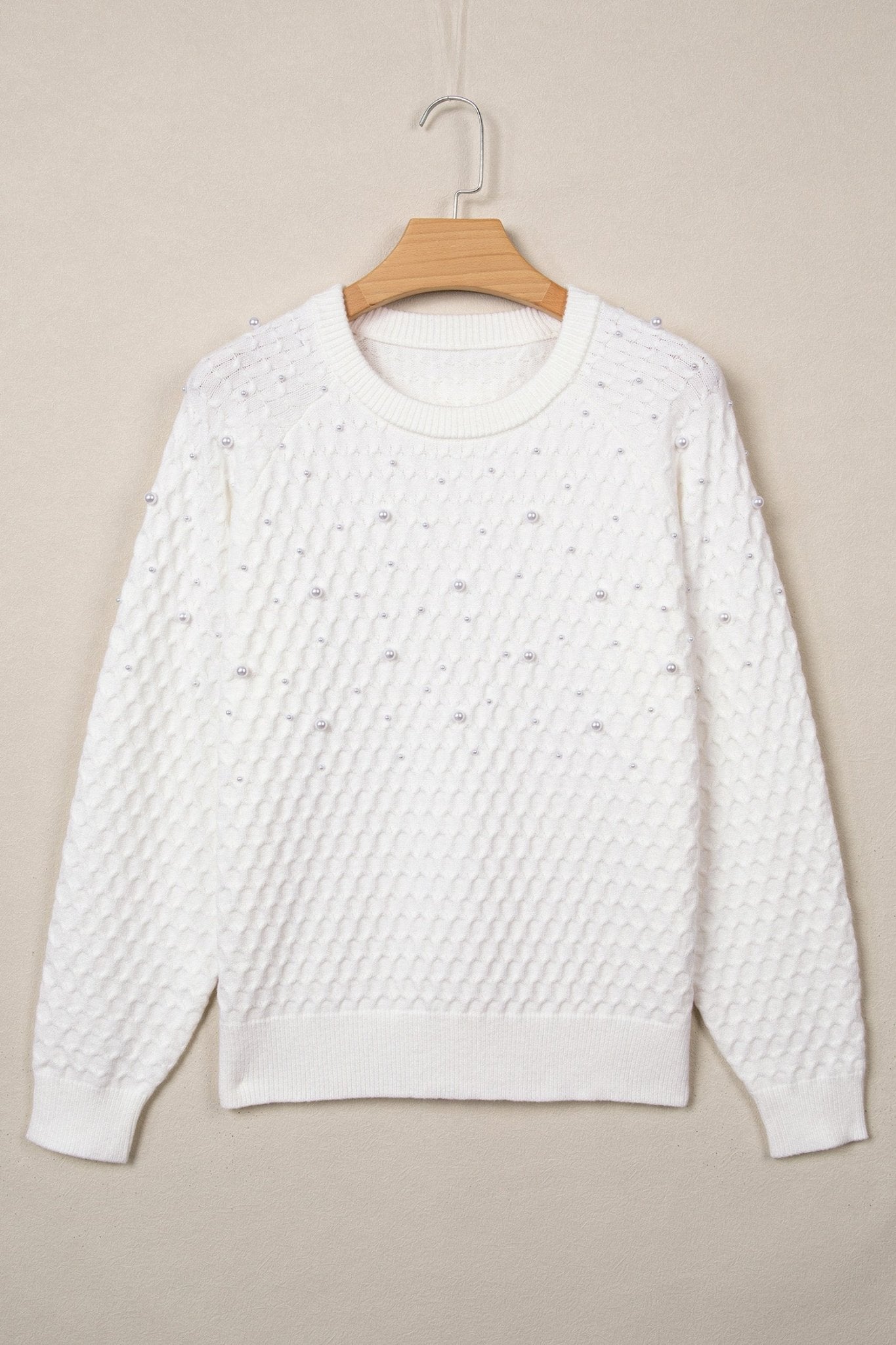 White textured knit crew-neck sweater hanging on a wooden hanger against a beige background.