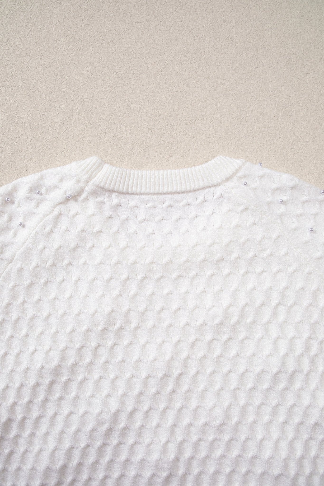 Close-up of a white textured knit sweater with a ribbed crew neck, laid flat against a light beige background.