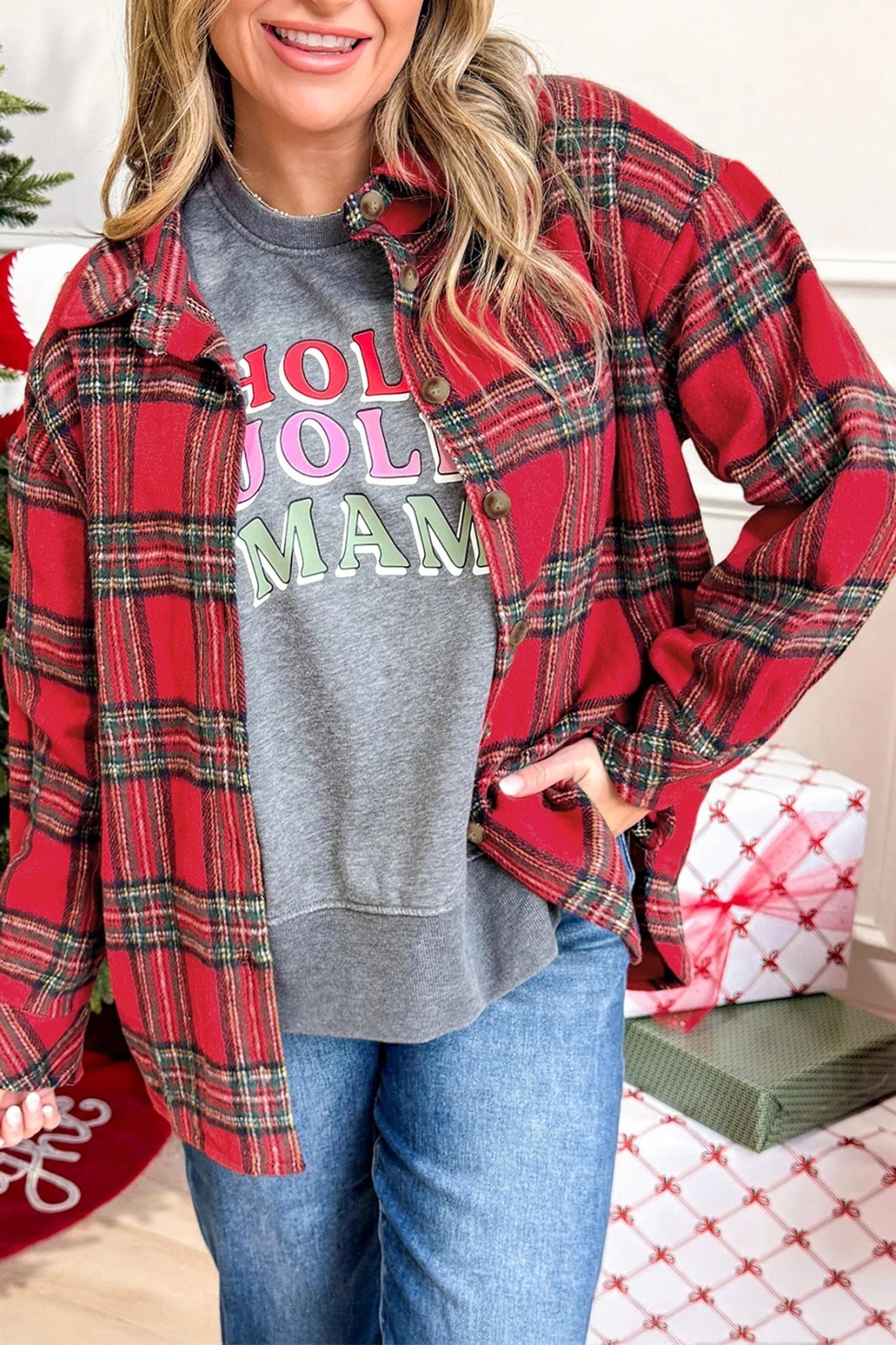 A woman wearing a red and green plaid flannel button-up shirt over a gray graphic sweatshirt that reads 'HOLY MOLY MAM' in pink and green letters, paired with blue jeans. The background shows a cozy holiday setting with wrapped gifts and a stocking.