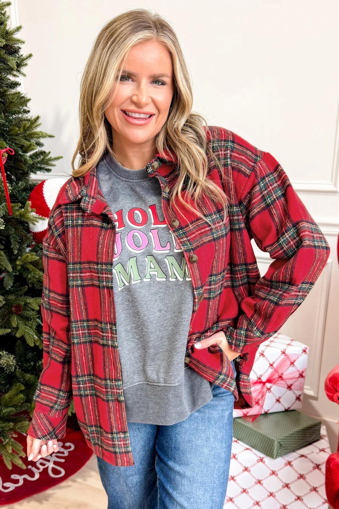 A woman wearing a red and green plaid flannel shirt over a gray graphic sweatshirt that reads 'HOLIDAY MAM' in colorful letters, paired with blue jeans. She is standing indoors next to a decorated Christmas tree with wrapped gifts on the floor.