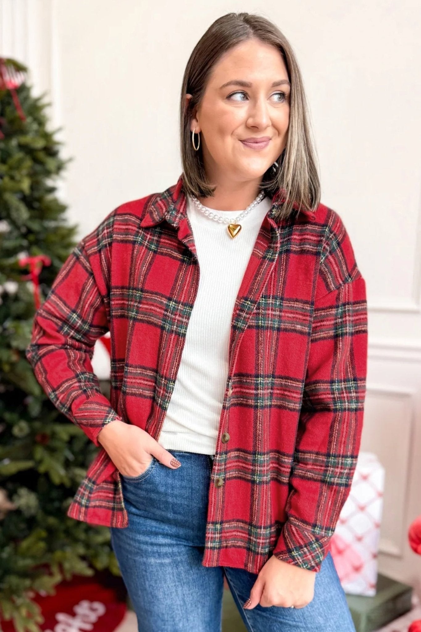 A woman wearing a red and black plaid button-up shirt over a white top and blue jeans, standing in front of a decorated Christmas tree.