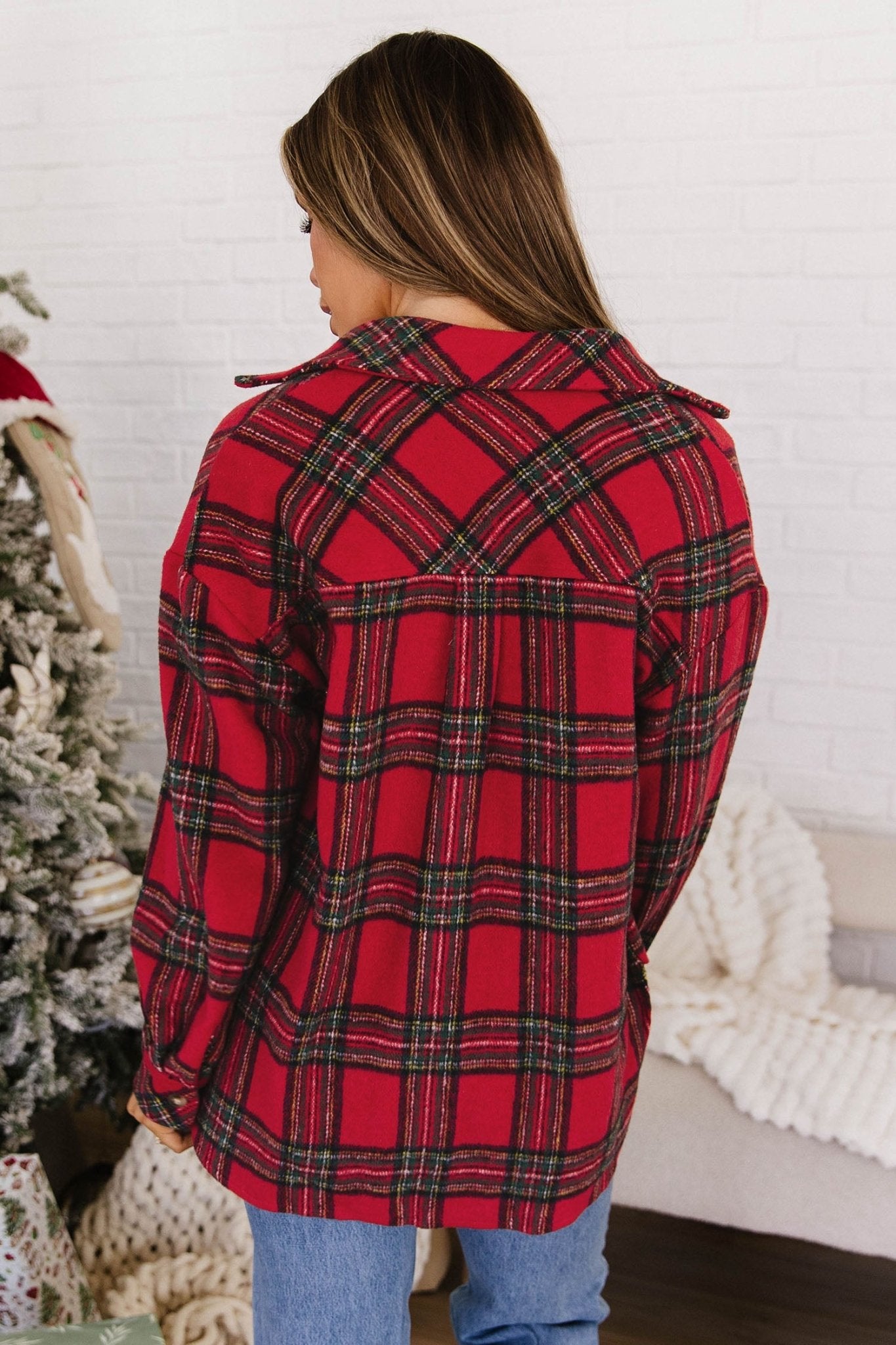 A woman shown from the back wearing a red plaid flannel shirt with black and green stripes, paired with blue jeans, standing in a cozy indoor setting with a white brick wall, a Christmas tree, and a white knit blanket.