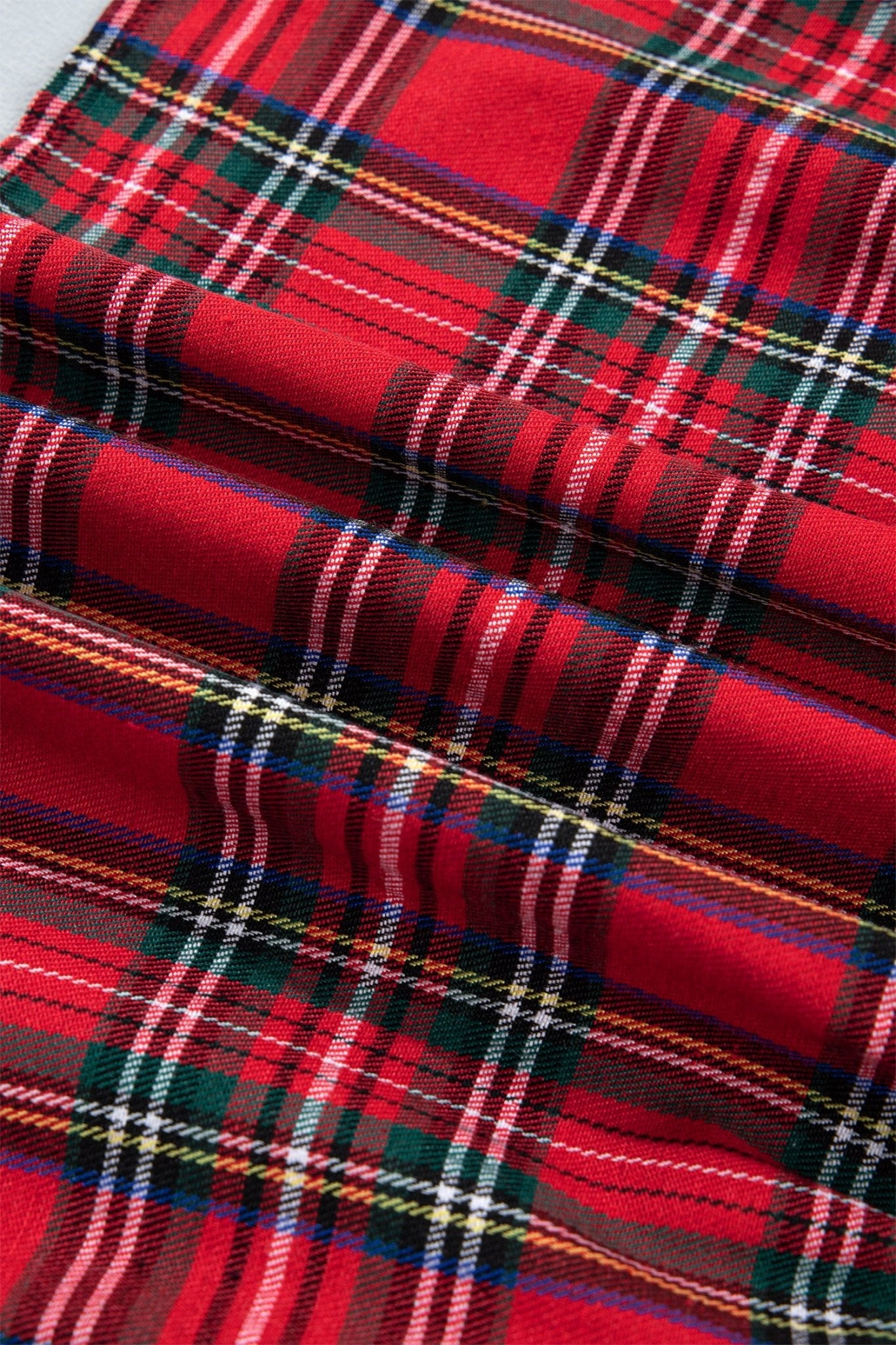 A close-up of a red tartan plaid fabric with green, white, and thin yellow stripes, shown in soft folds.