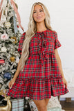 Woman wearing a red and green plaid short-sleeve dress with ruffled tie neckline by a Christmas tree.