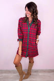 A woman modeling a red and green plaid mini shirt dress with brown suede boots, standing against a light pink wall.