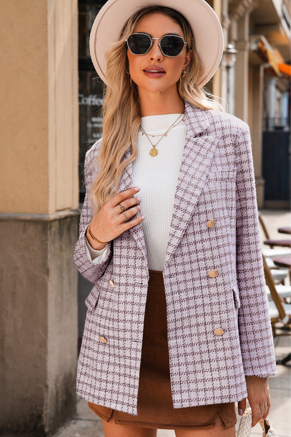 Woman wearing a plaid blazer, white top, and brown skirt with sunglasses and a hat.