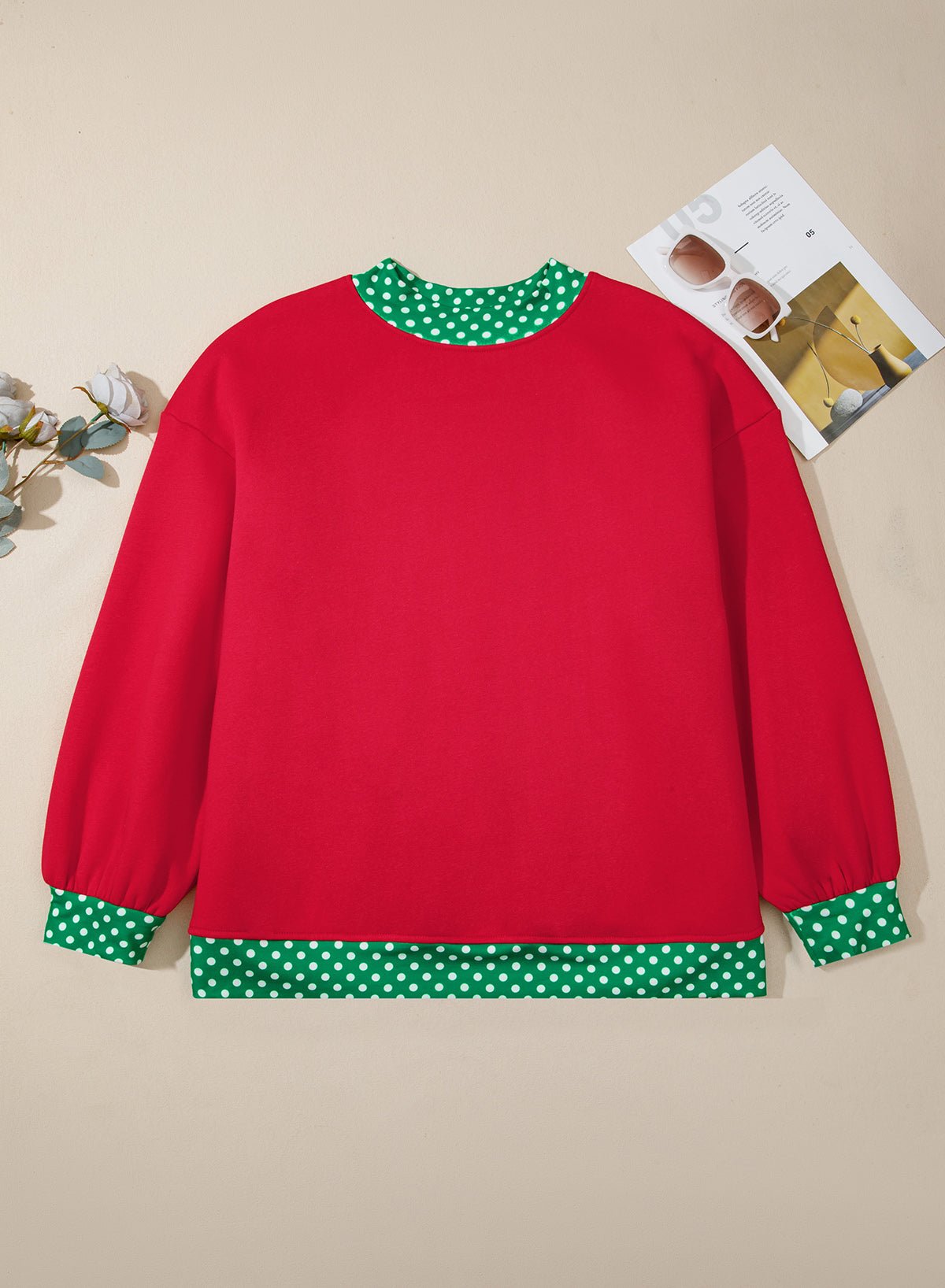 Red long-sleeve sweatshirt with green-white polka-dot trim, displayed flat on beige with magazine and flowers