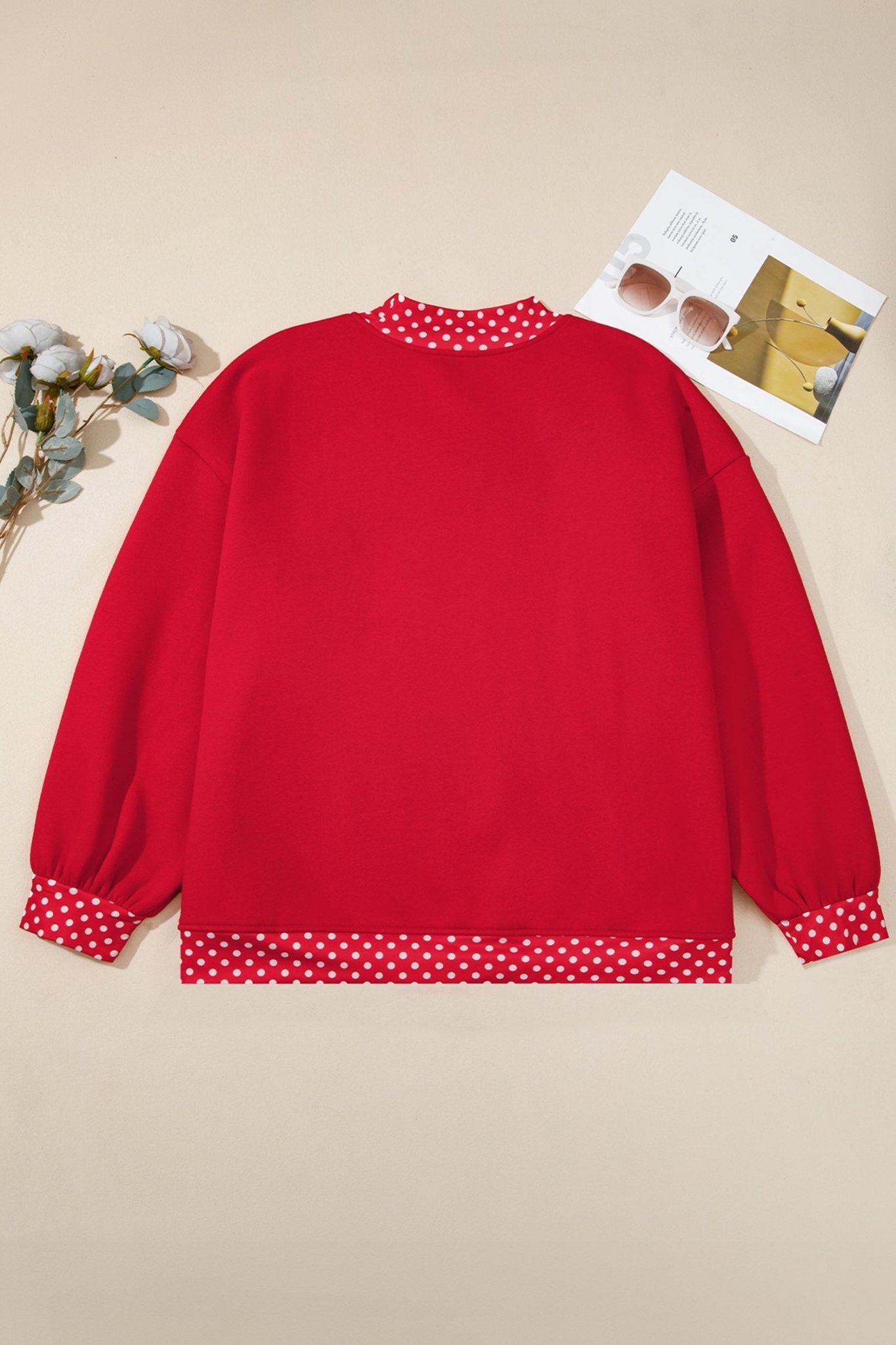 Red long-sleeve sweatshirt with red-white polka-dot trim, laid flat on beige with flowers and a magazine.