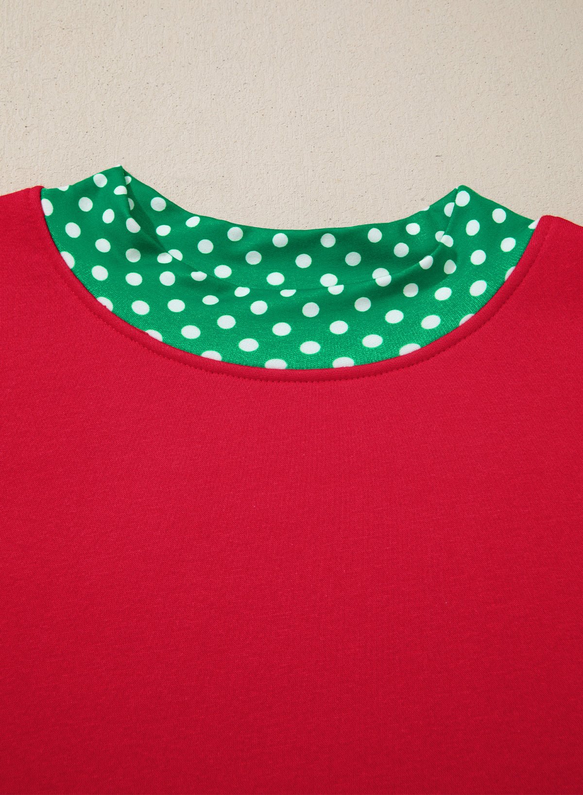 Close-up of a red garment with a contrasting green and white polka dot inner collar, shown against a plain beige background.