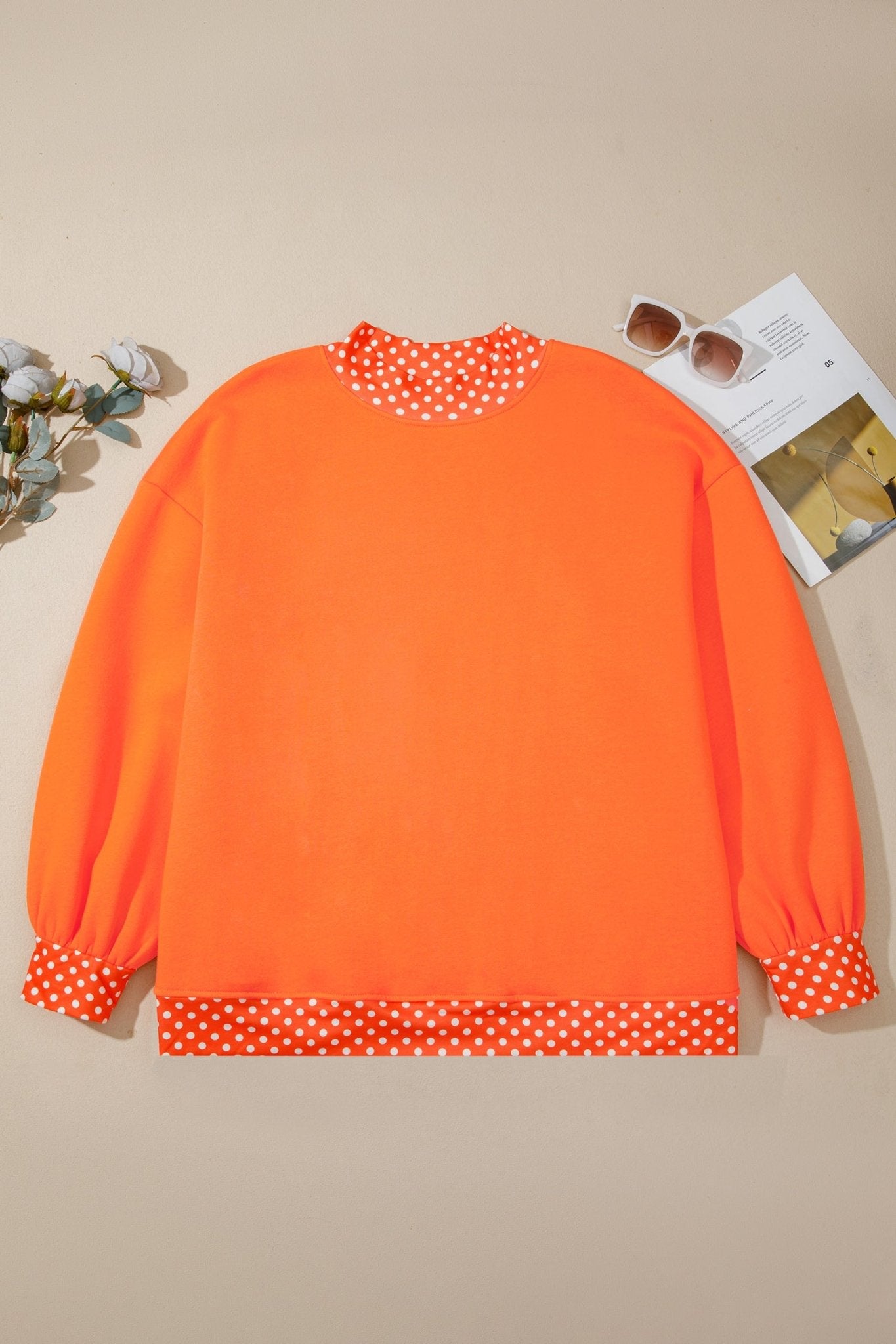 Bright orange sweatshirt with white polka-dot trim, styled flat on beige with sunglasses and flowers.