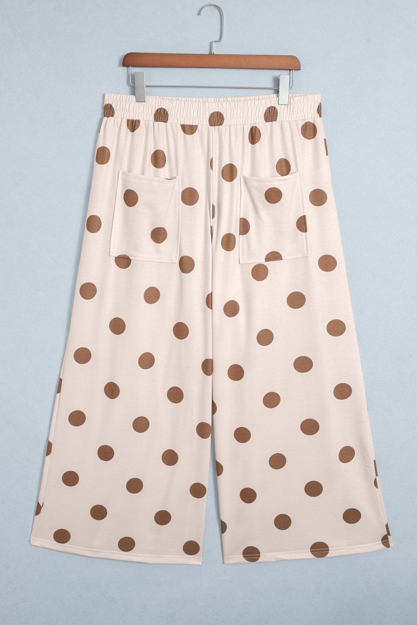 Cream wide-leg cropped pants with brown polka dots, hanging on a wooden hanger against light blue-gray.
