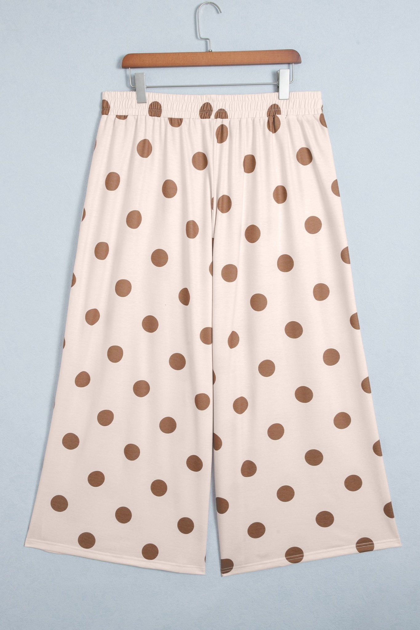 Cream wide-leg cropped pants with brown polka dots, hanging on a wooden hanger against light blue-gray.