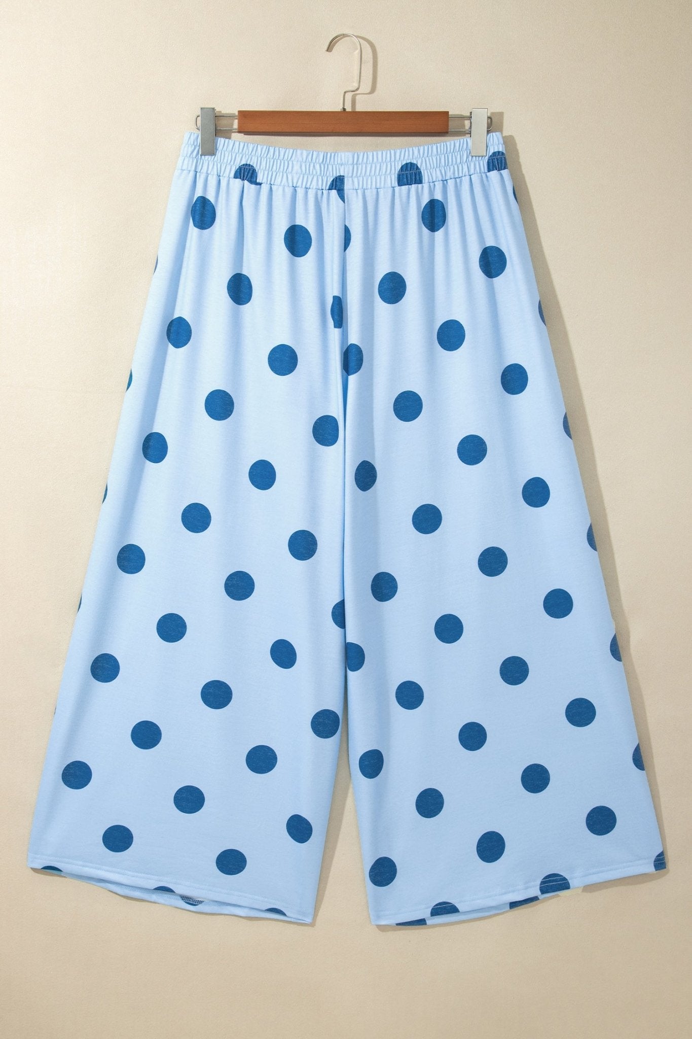Light blue cropped pants with dark blue polka dots, hanging on a wooden hanger against a beige wall.