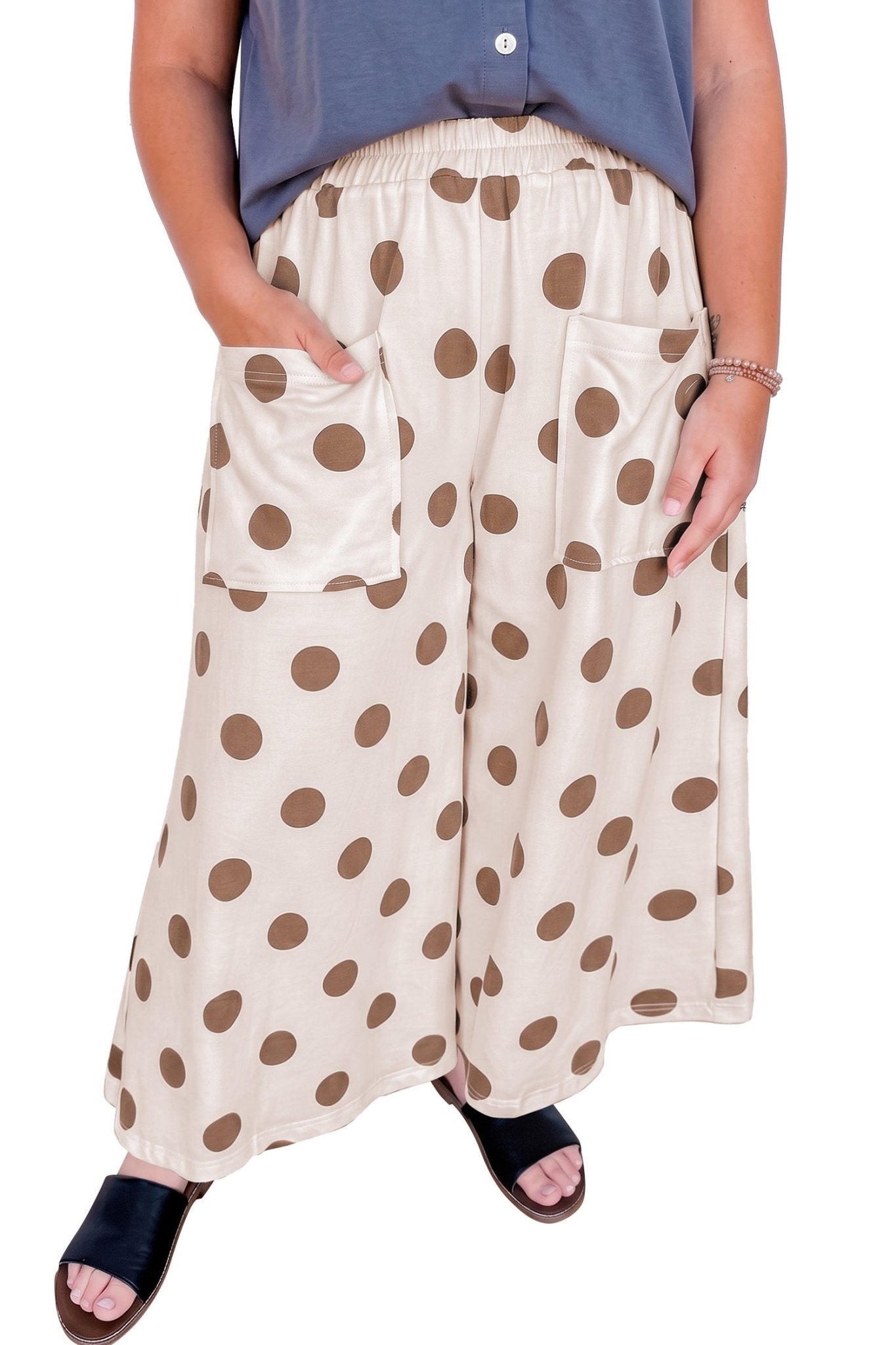 Person in cream wide-leg brown polka dot pants, blue sleeveless top, and black slides on white.