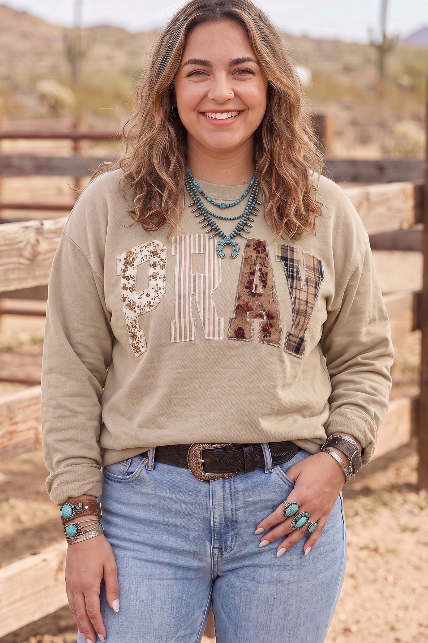 Woman in beige “PRAY” embellished sweatshirt with blue jeans and turquoise jewelry.