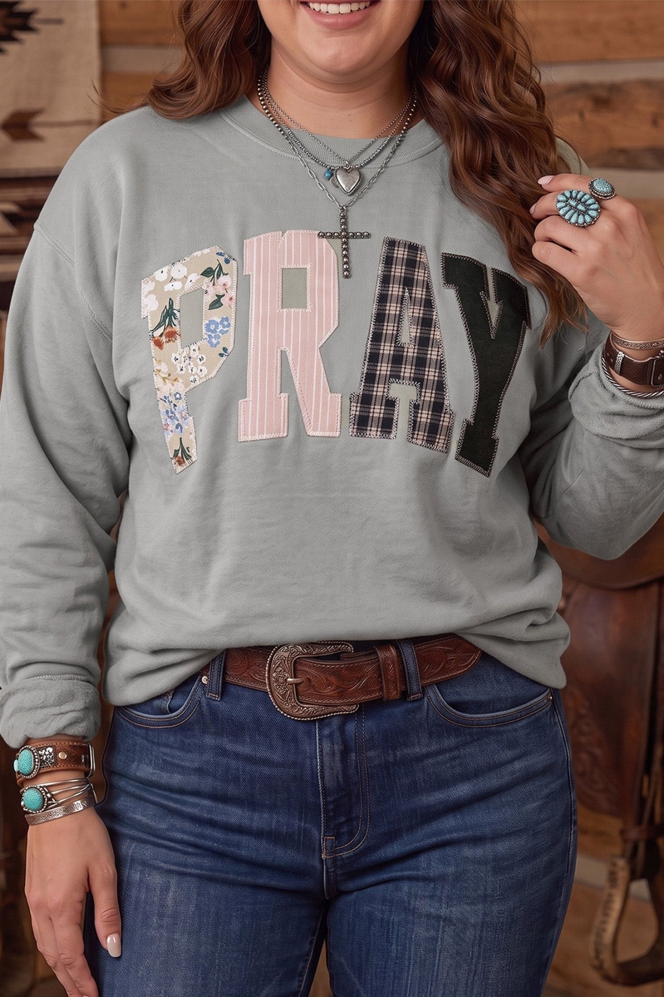 Woman in gray “PRAY” patterned sweatshirt with blue jeans and western jewelry.