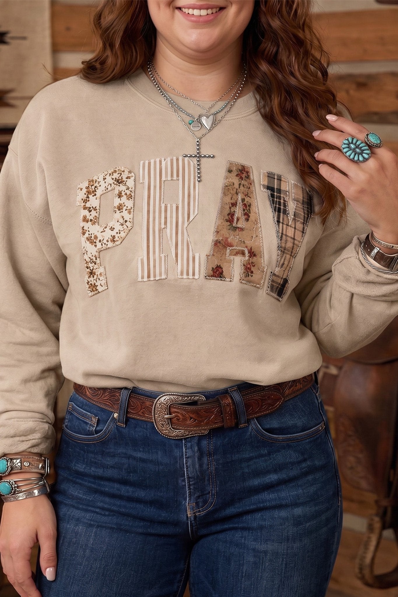 Woman in beige “PRAY” patterned sweatshirt with blue jeans and brown belt.