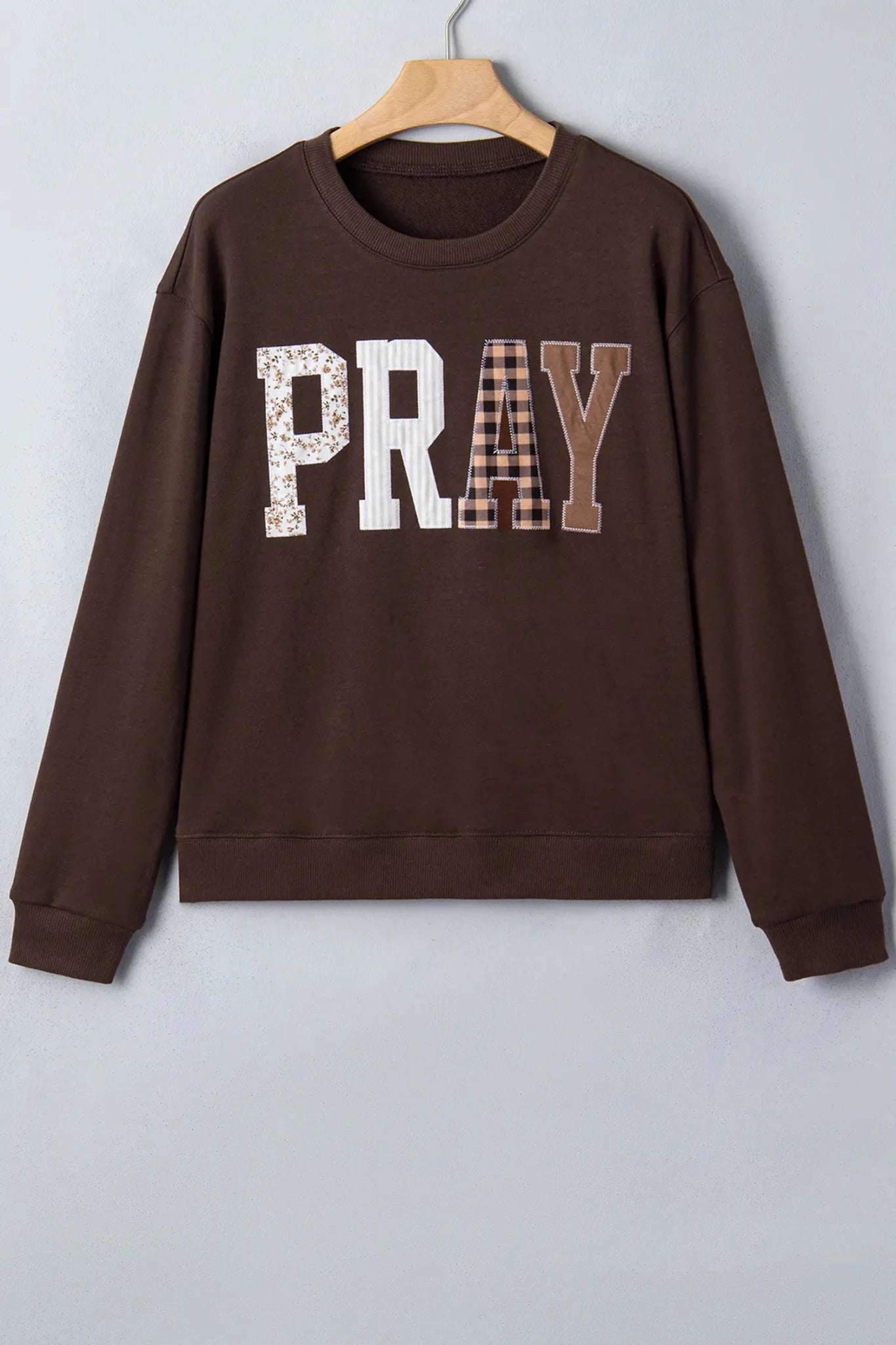 Brown “PRAY” sweatshirt with floral, gingham, and solid letter patterns on hanger.