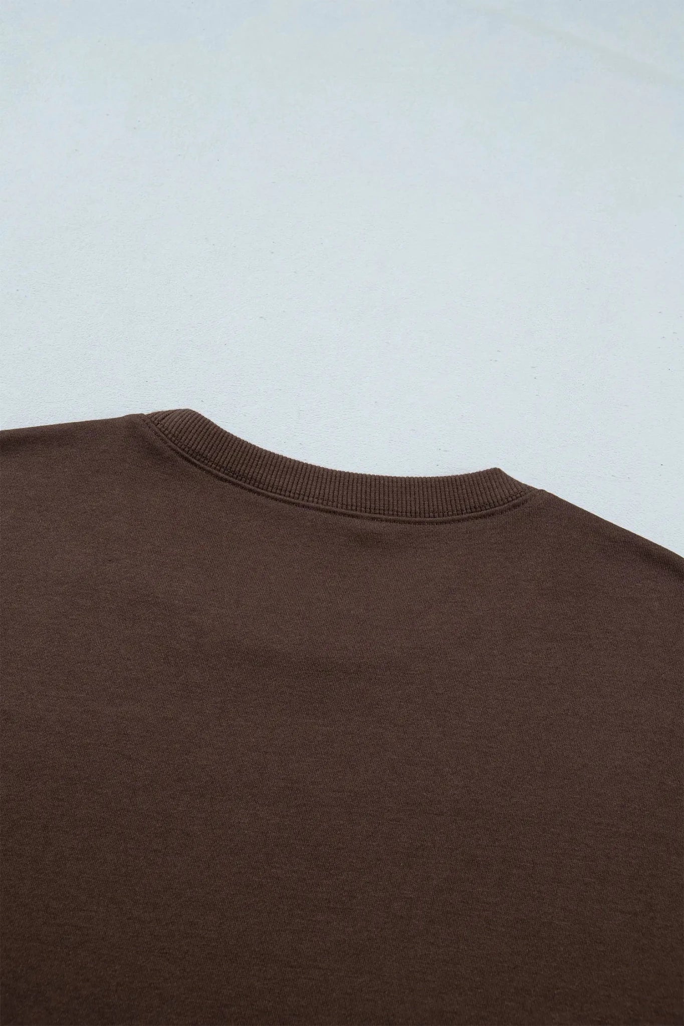 Close-up of a brown crewneck sweatshirt showing the ribbed collar against a light gray background.