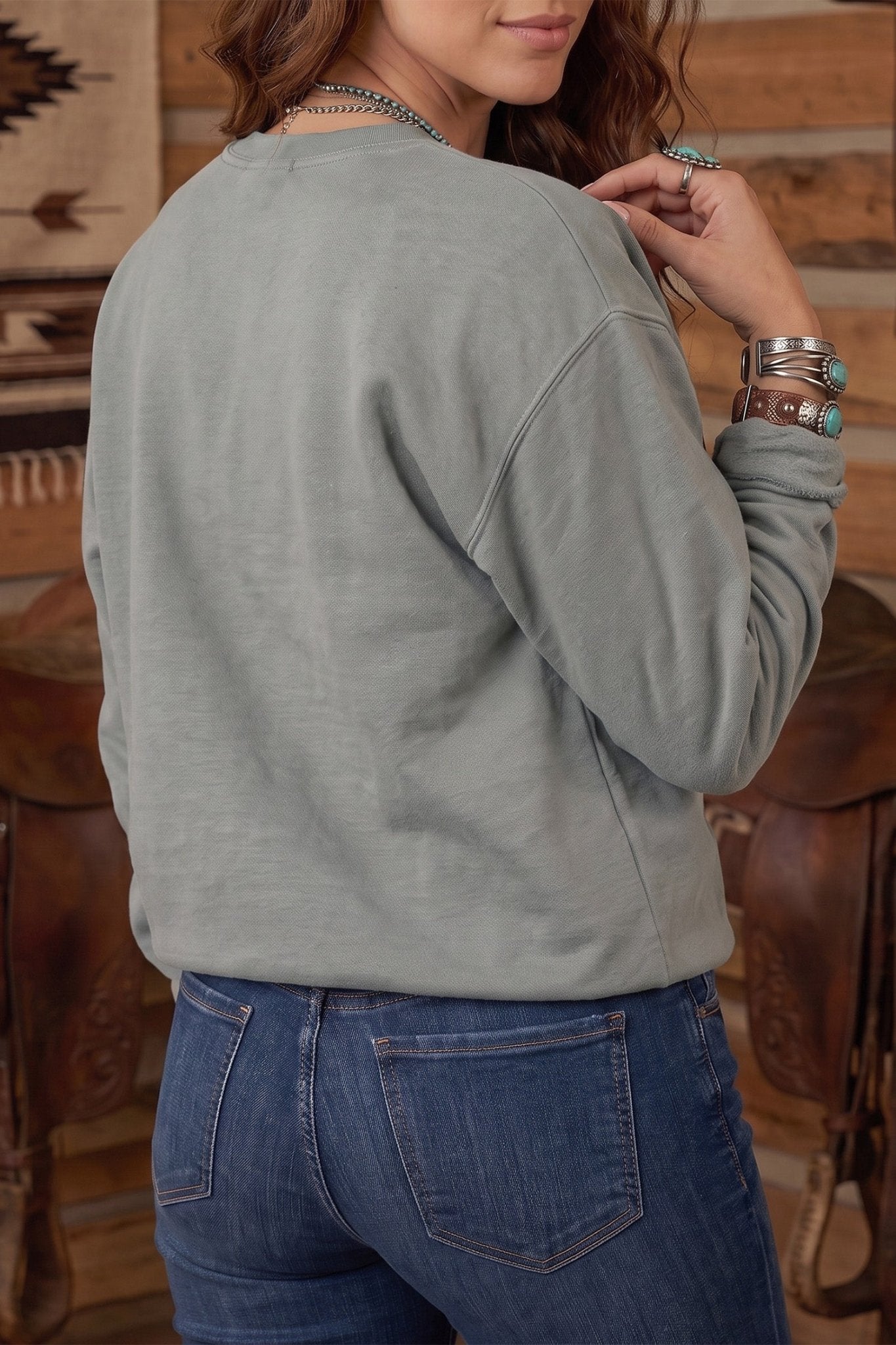Woman in sage green crewneck sweatshirt and blue jeans in rustic western setting.