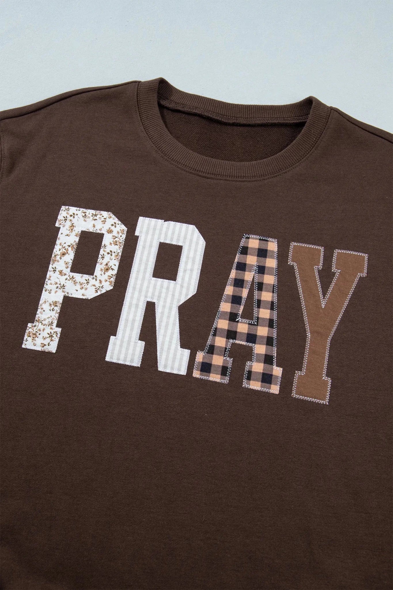 Close-up of brown “PRAY” tee with floral, striped, gingham, and solid letter patterns.
