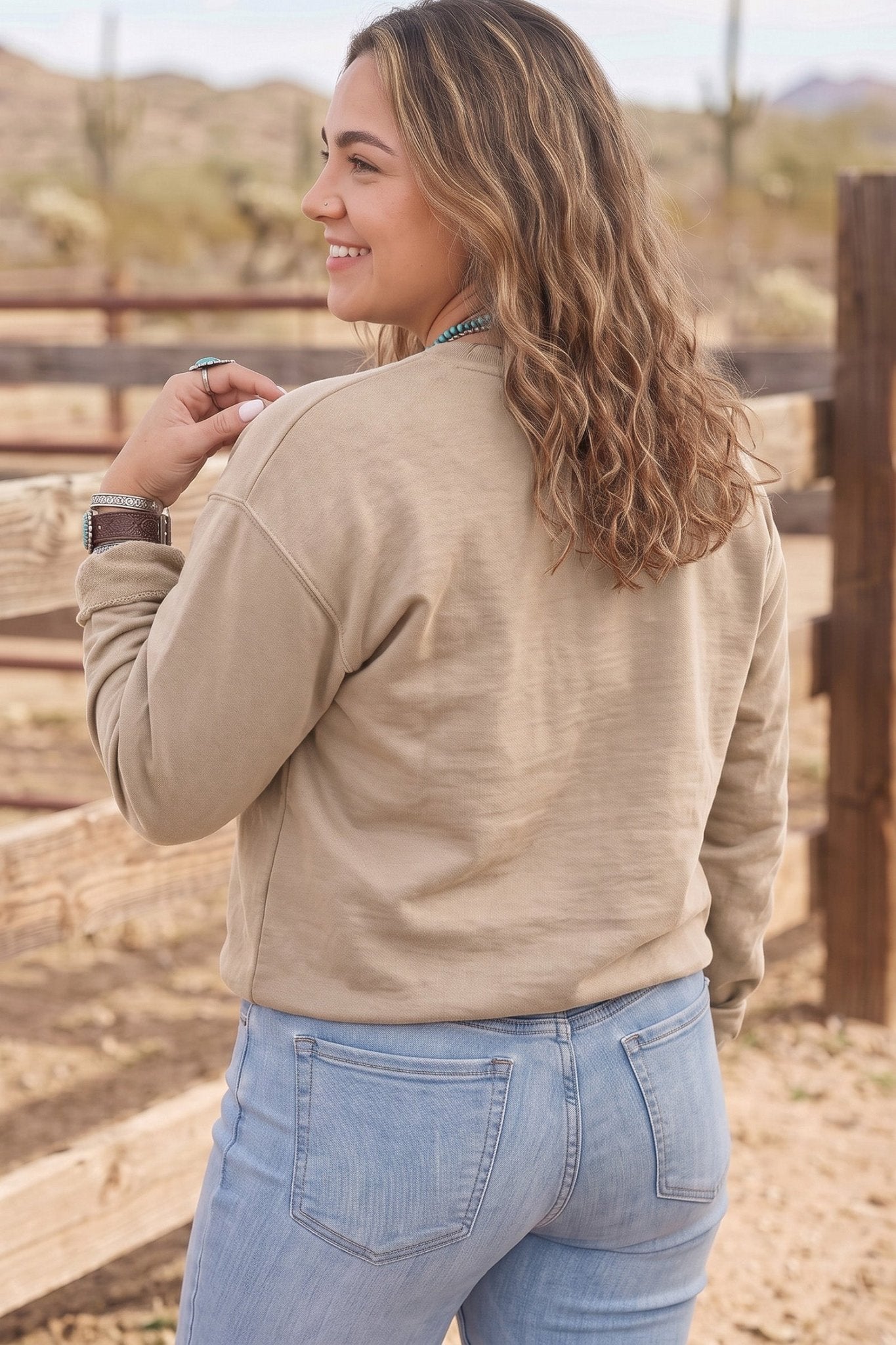 Back view of woman in beige sweatshirt and light blue jeans near wooden fence.
